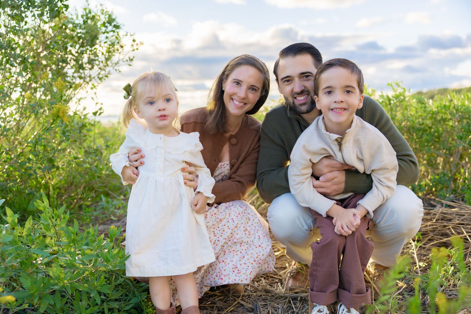 A smiling family of four with two parents, a young boy and a toddler girl in a white dress posing outdoors in a grassy field at sunset.