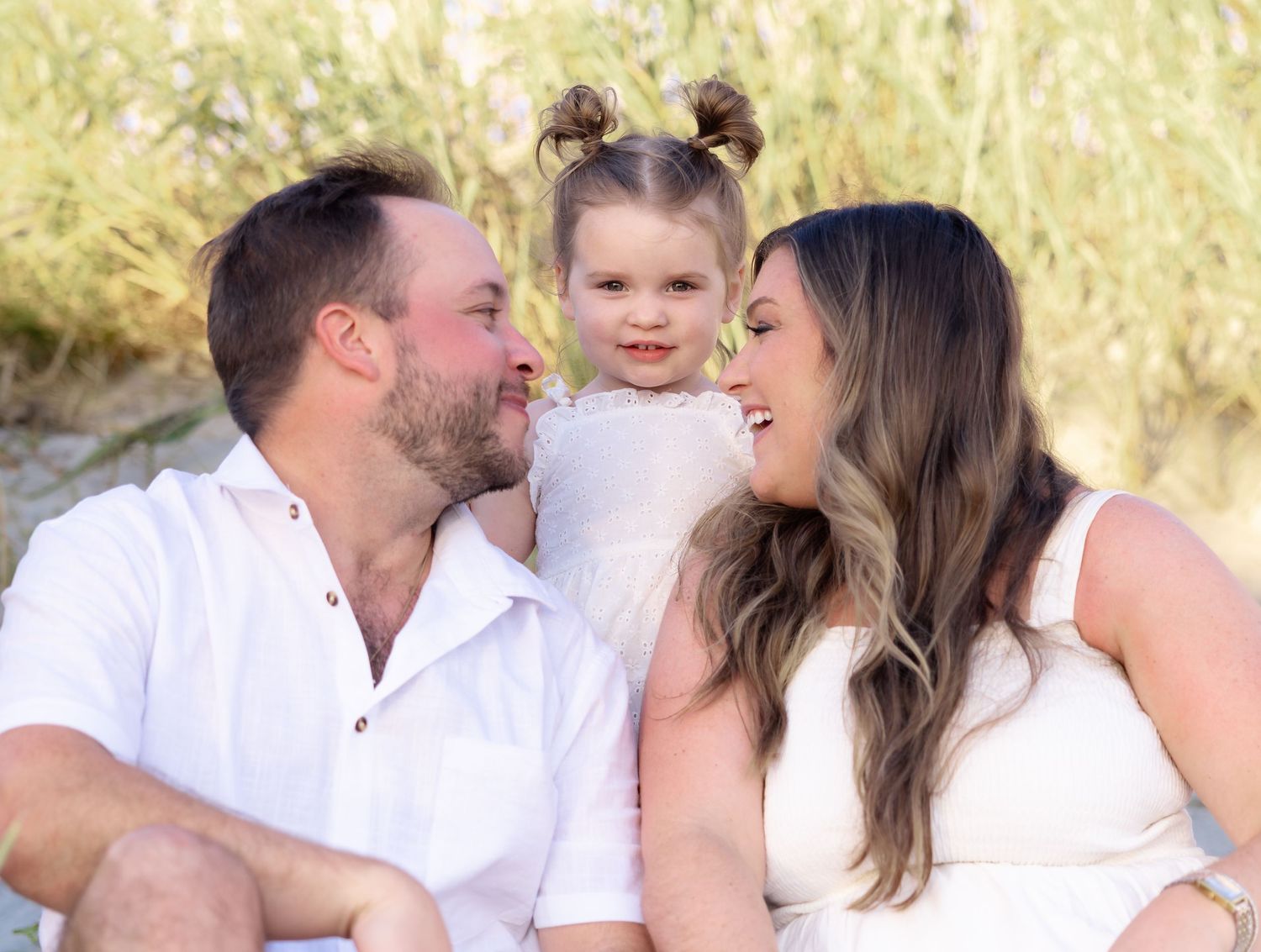 A smiling man in a white shirt and woman in a white dress sit beside a young girl with pigtail buns during an outdoor family photo.