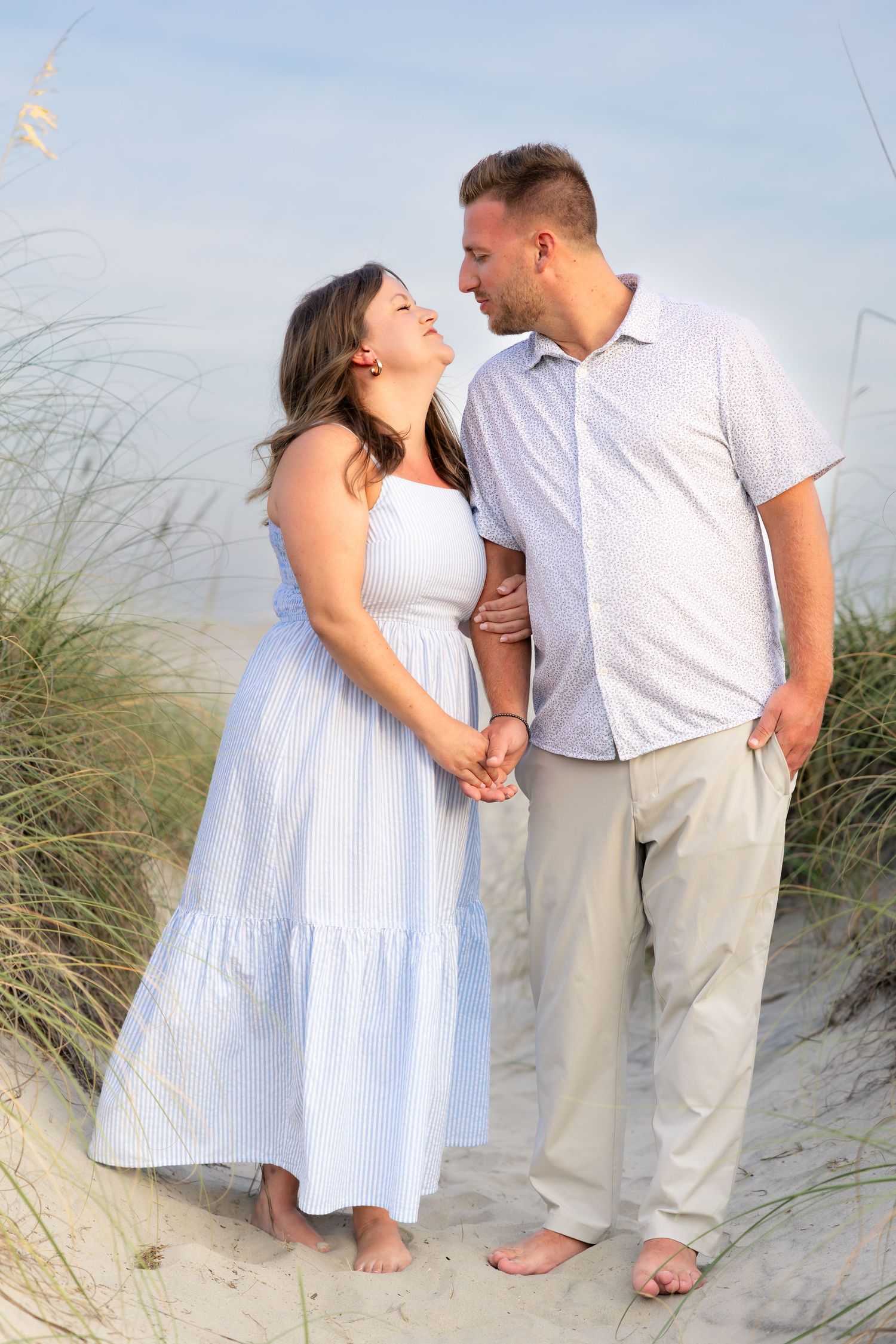 Couple holding hands on sandy beach dunes, woman in white dress and man in light shirt, sharing a romantic moment.