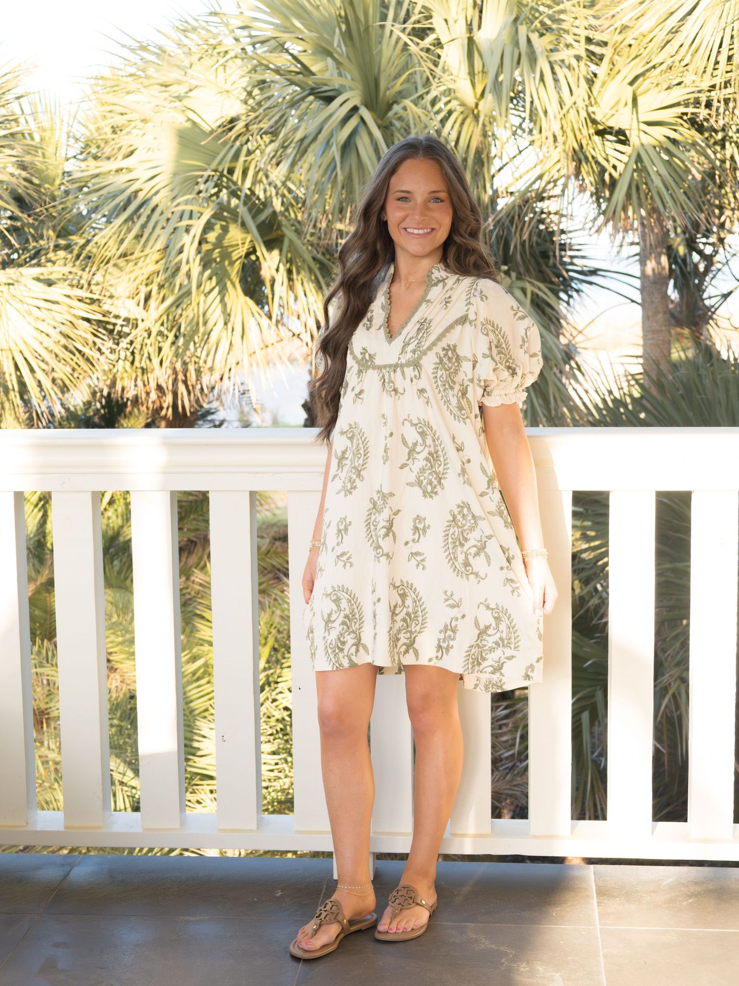 A young woman with long dark hair wearing a floral print dress stands on a wooden deck with palm trees in the background.