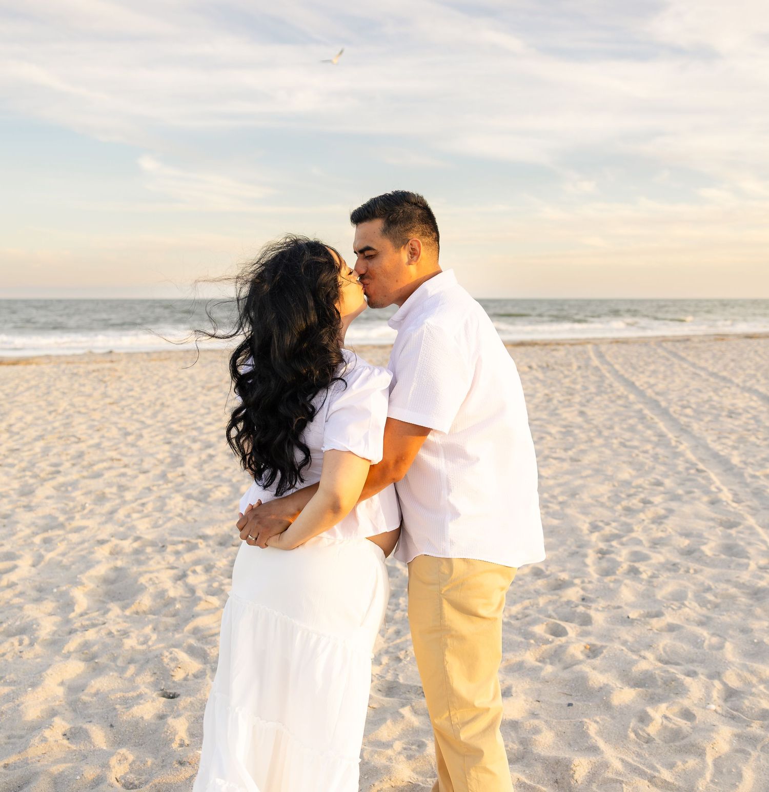 Couple kissing on a sandy beach at sunset, both dressed in white and tan outfits with ocean waves in the background.