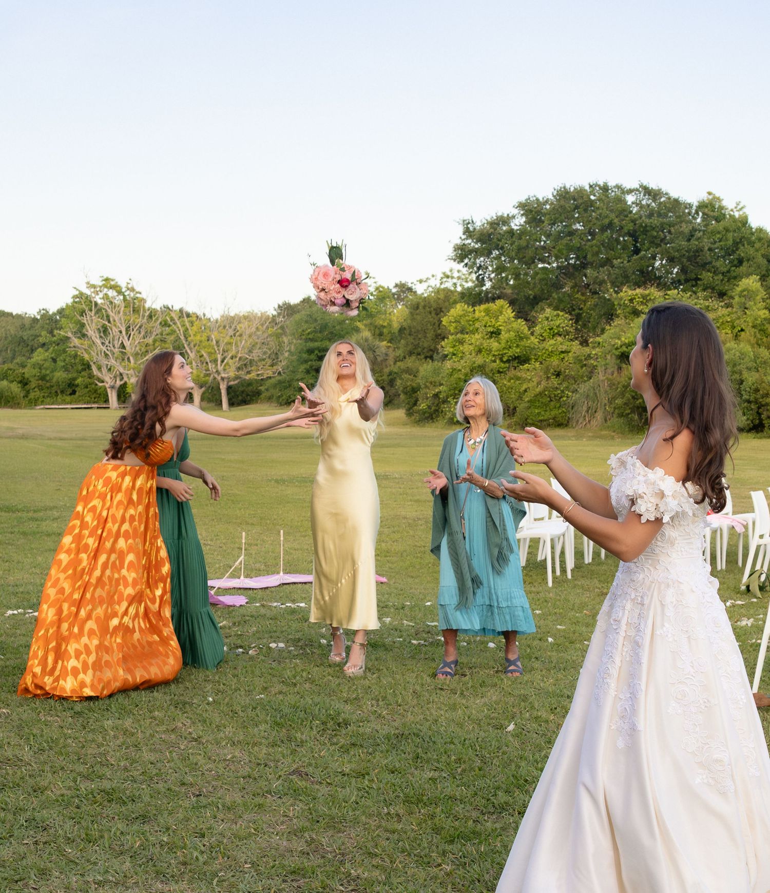 Bride in lace gown tosses pink bouquet to three women in colorful dresses on green lawn at wedding.