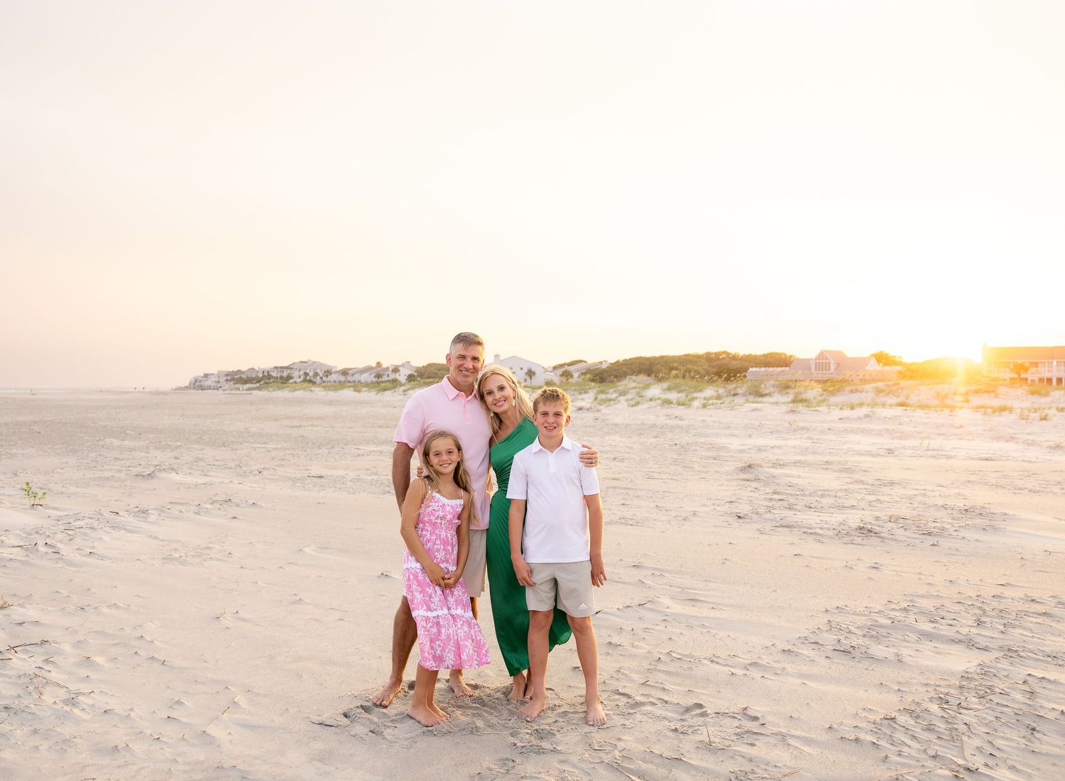 Family of four posing together on a sandy beach at golden sunset hour near coastal homes.