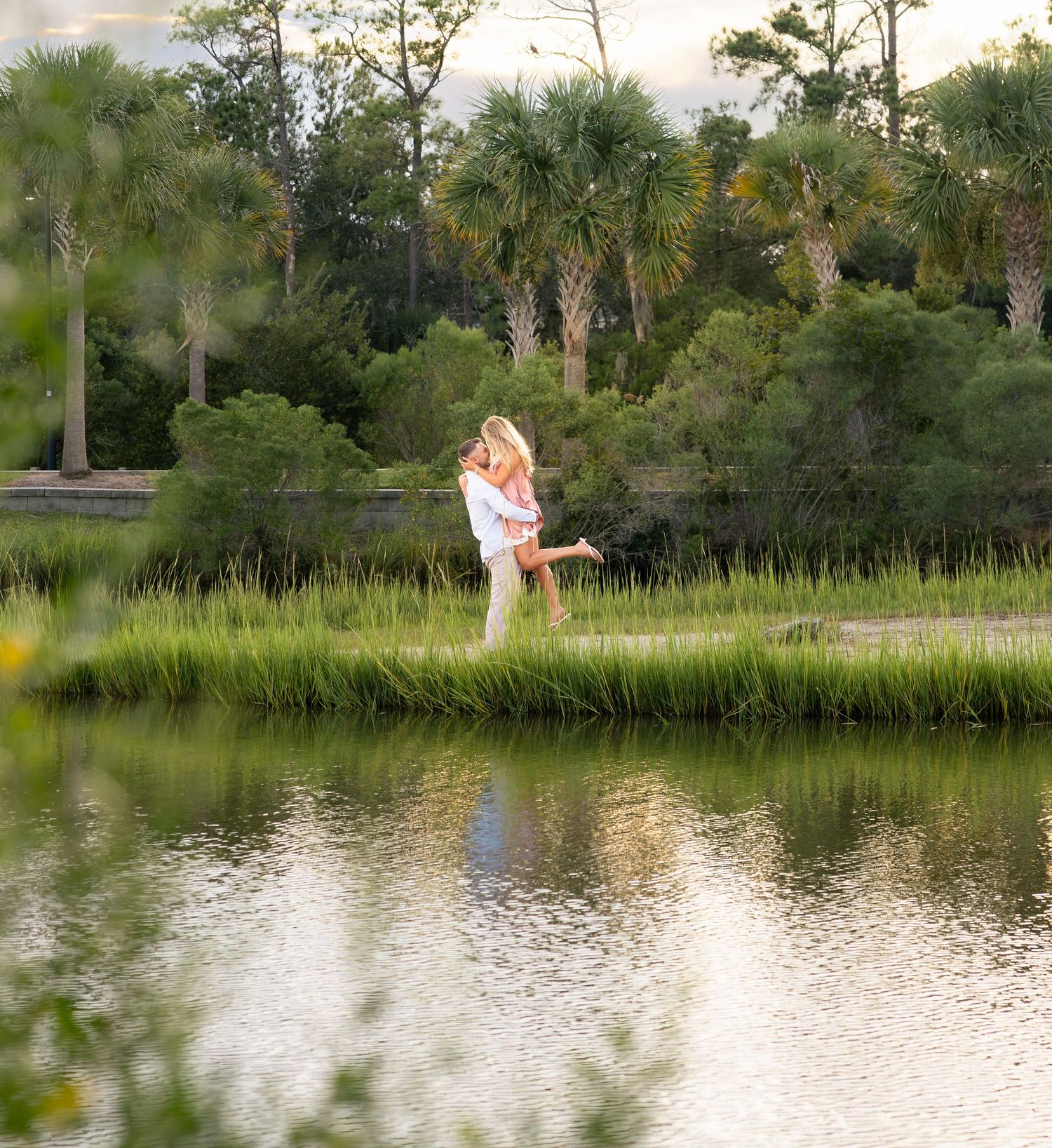 Couple embracing by a serene pond surrounded by lush green marsh grass and palm trees at golden hour.