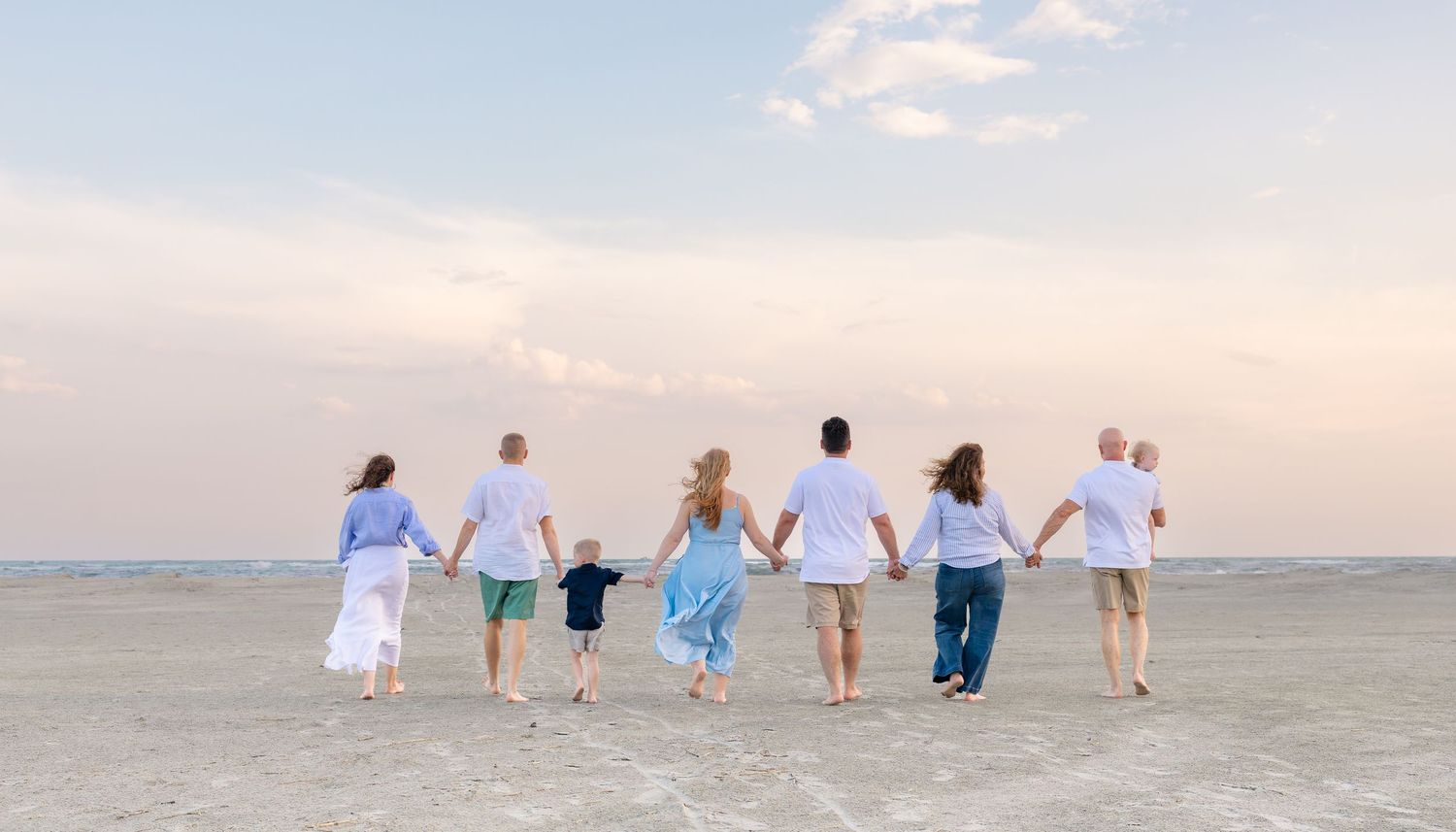 Family of seven holding hands walking on a sandy beach at sunset with pastel sky backdrop.
