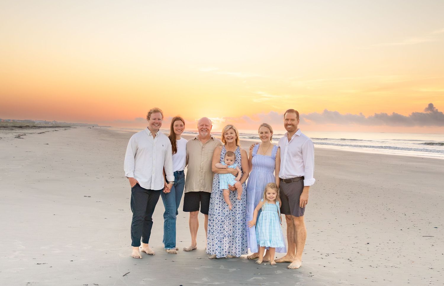 Family of seven posing on a sandy beach at golden sunset, wearing coordinated outfits in blue and white tones.