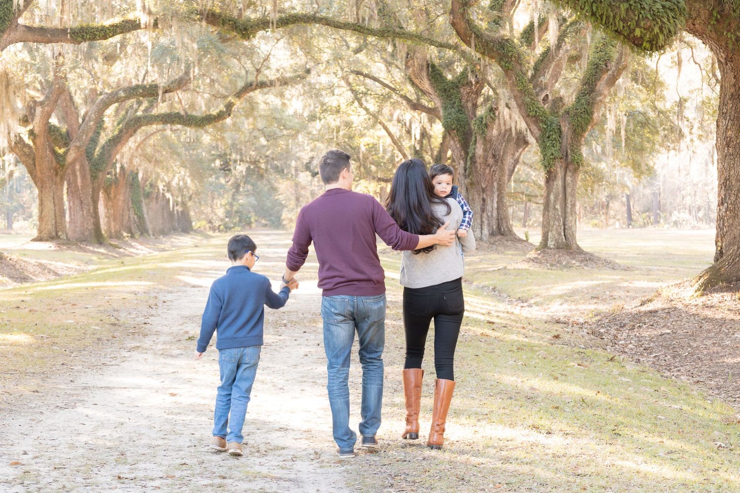 Family of four walking hand-in-hand down a scenic tree-lined path with Spanish moss in warm golden light.