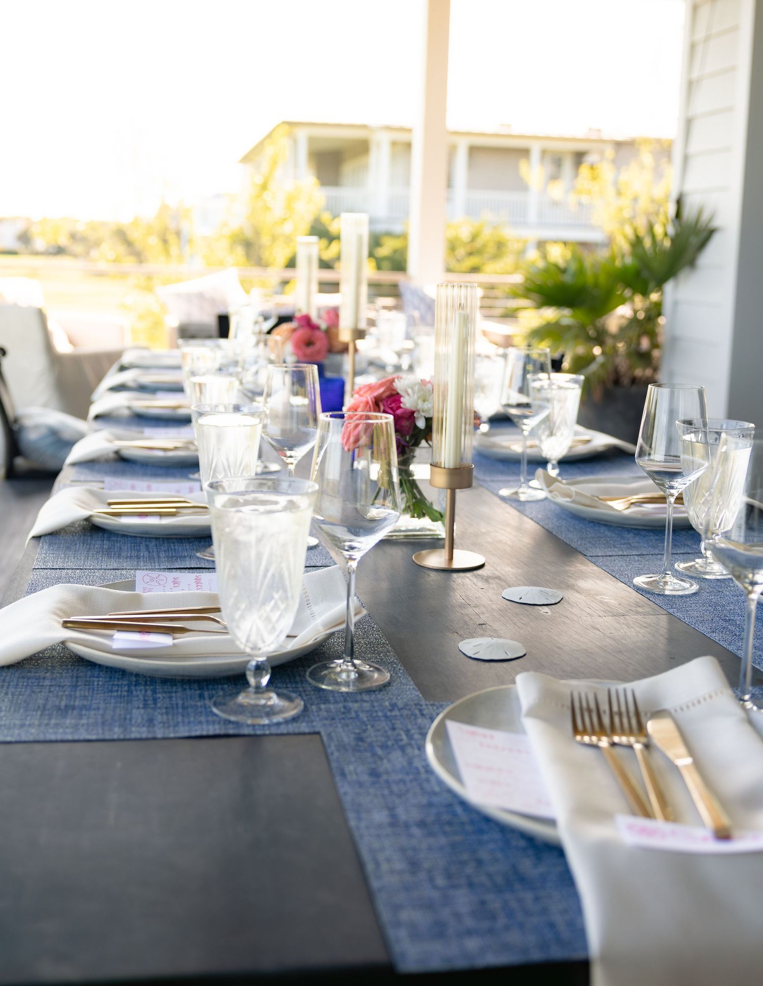 Elegantly set outdoor dining table with blue runners, crystal glasses, and gold cutlery.