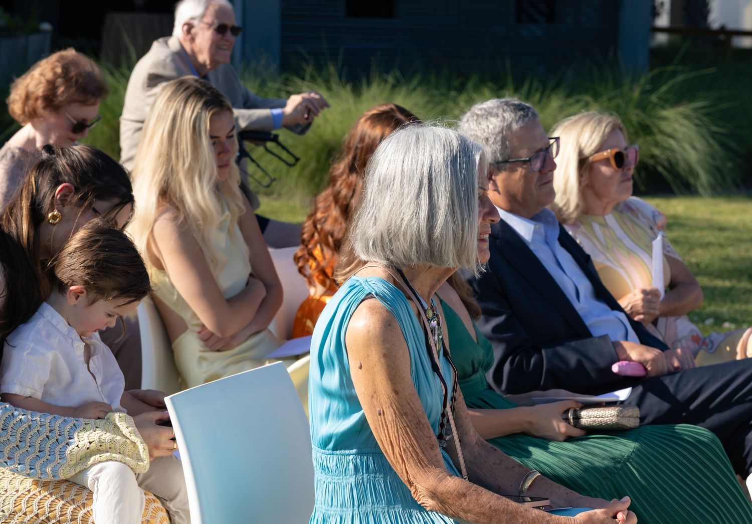 Group of elegantly dressed guests seated outdoors at a daytime event, including women with gray and blonde hair.