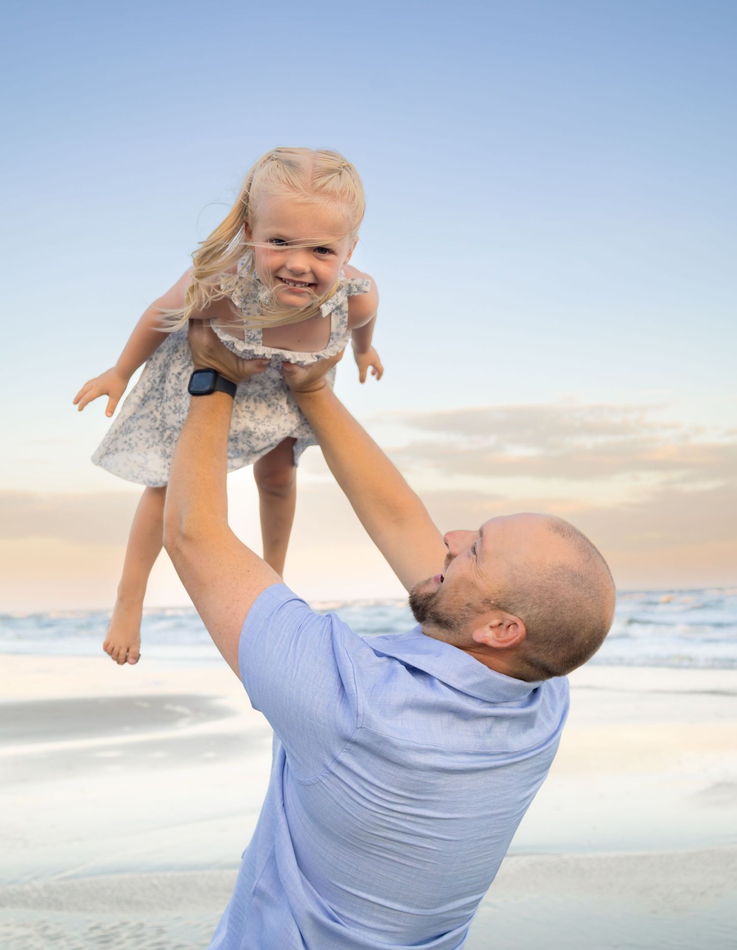 A bald father joyfully lifts his smiling toddler daughter in the air on a beach at sunset.