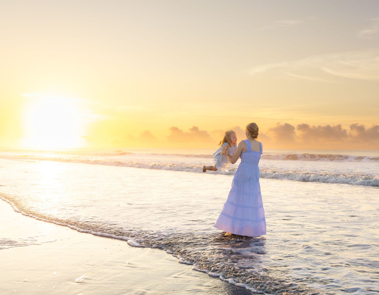 Woman in white dress holds child at golden sunset on beach shoreline with waves.