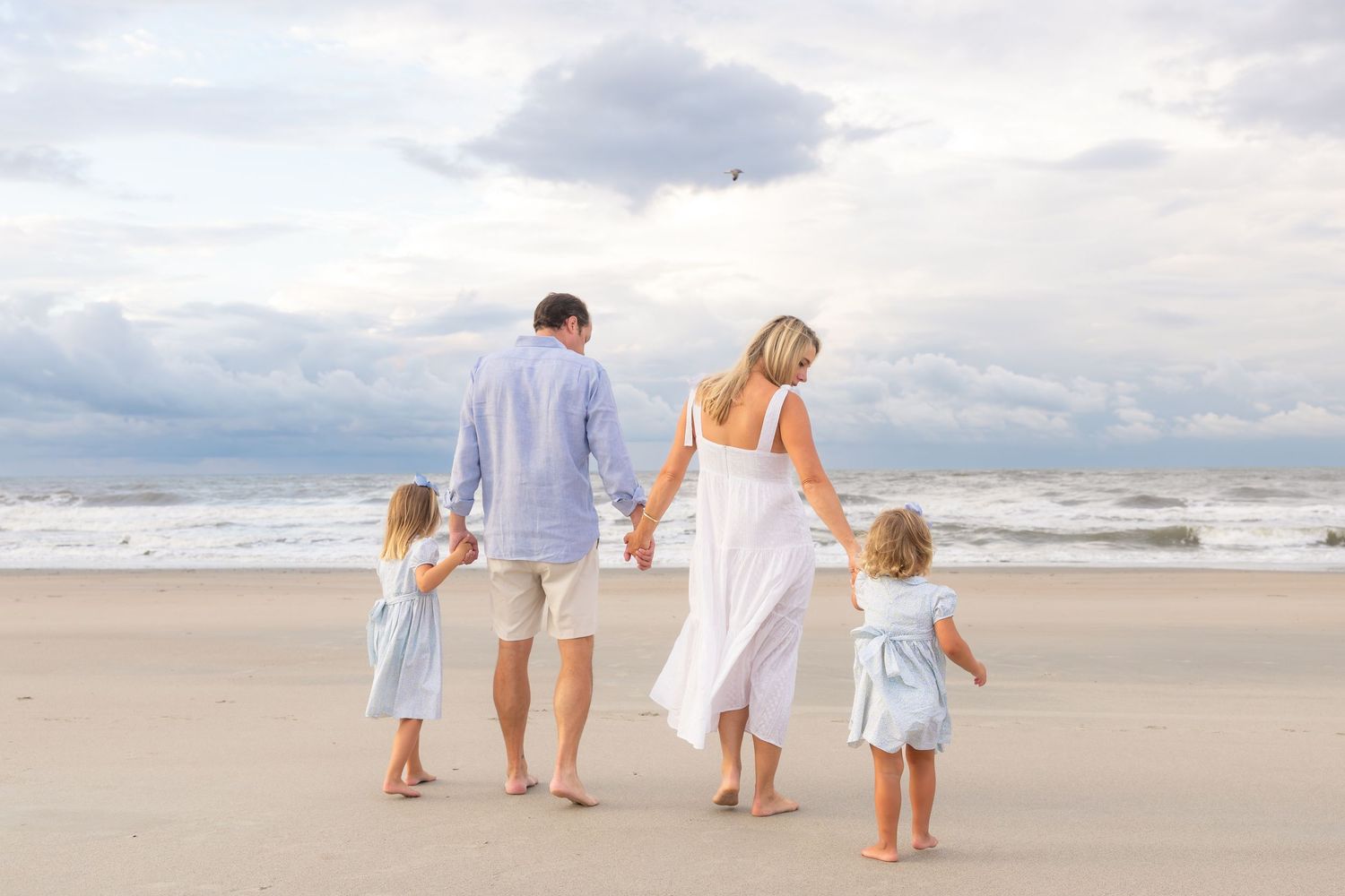 Family of four walking hand-in-hand on a sandy beach toward ocean waves under a cloudy sky.