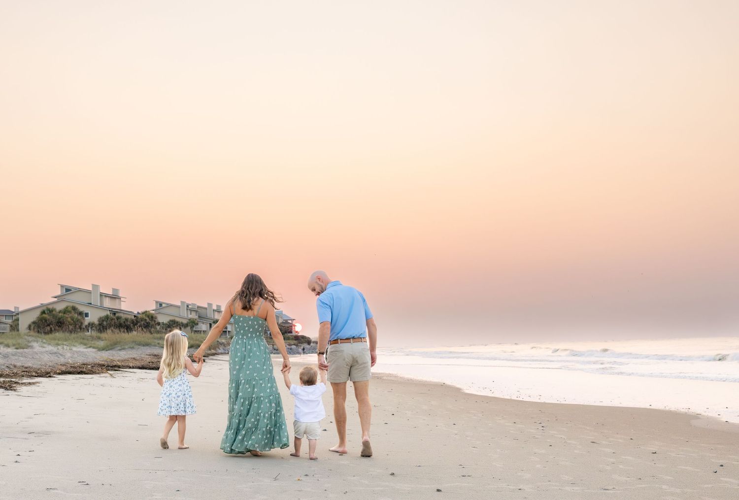 Family of four walking hand-in-hand on a beach at sunset, with pastel skies and ocean waves in the background.