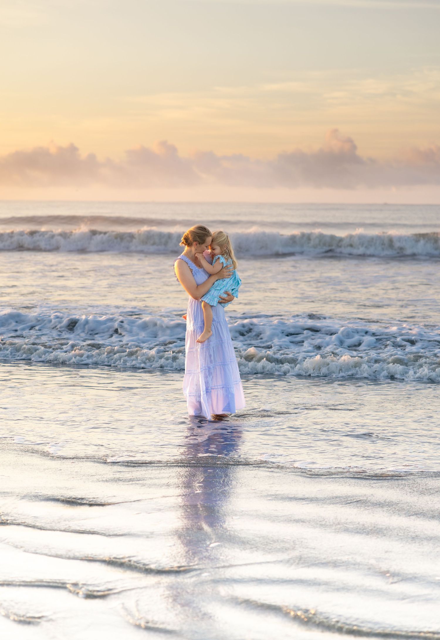 Mother holding young child at sunset beach, waves crashing behind them, both wearing light blue outfits.