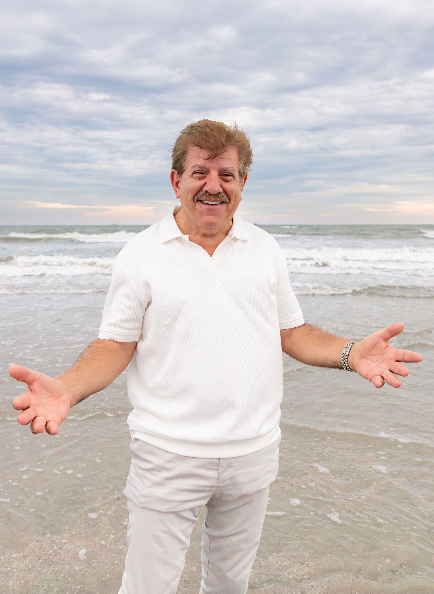 Smiling man in white polo shirt spreading arms wide on a sunny beach with ocean waves.