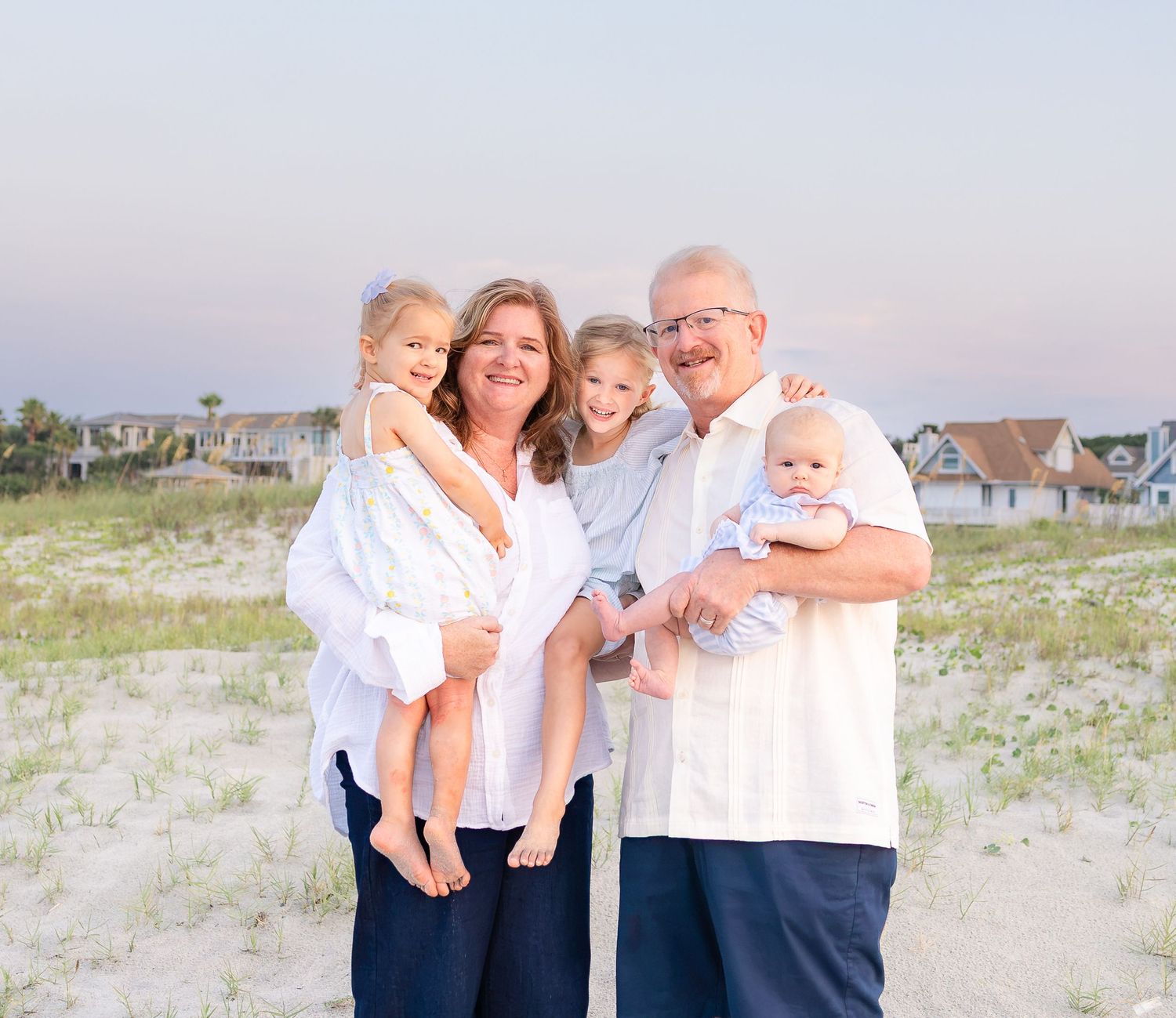 Family of five posing on a sandy beach at sunset, parents holding three young children, wearing white and navy outfits.