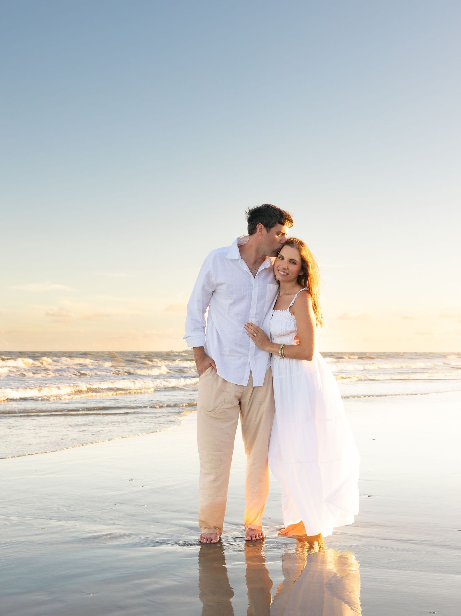 Couple in white outfits embracing on a beach at sunset with ocean waves in the background.