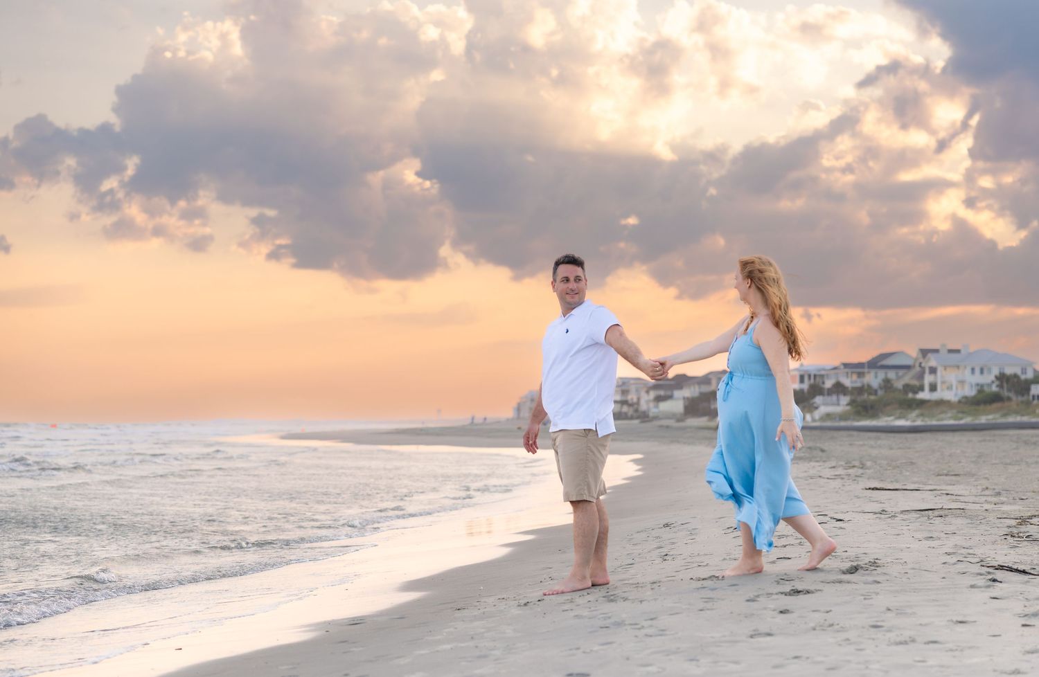 Couple holding hands walking on beach at sunset, woman in blue dress, romantic seaside stroll.