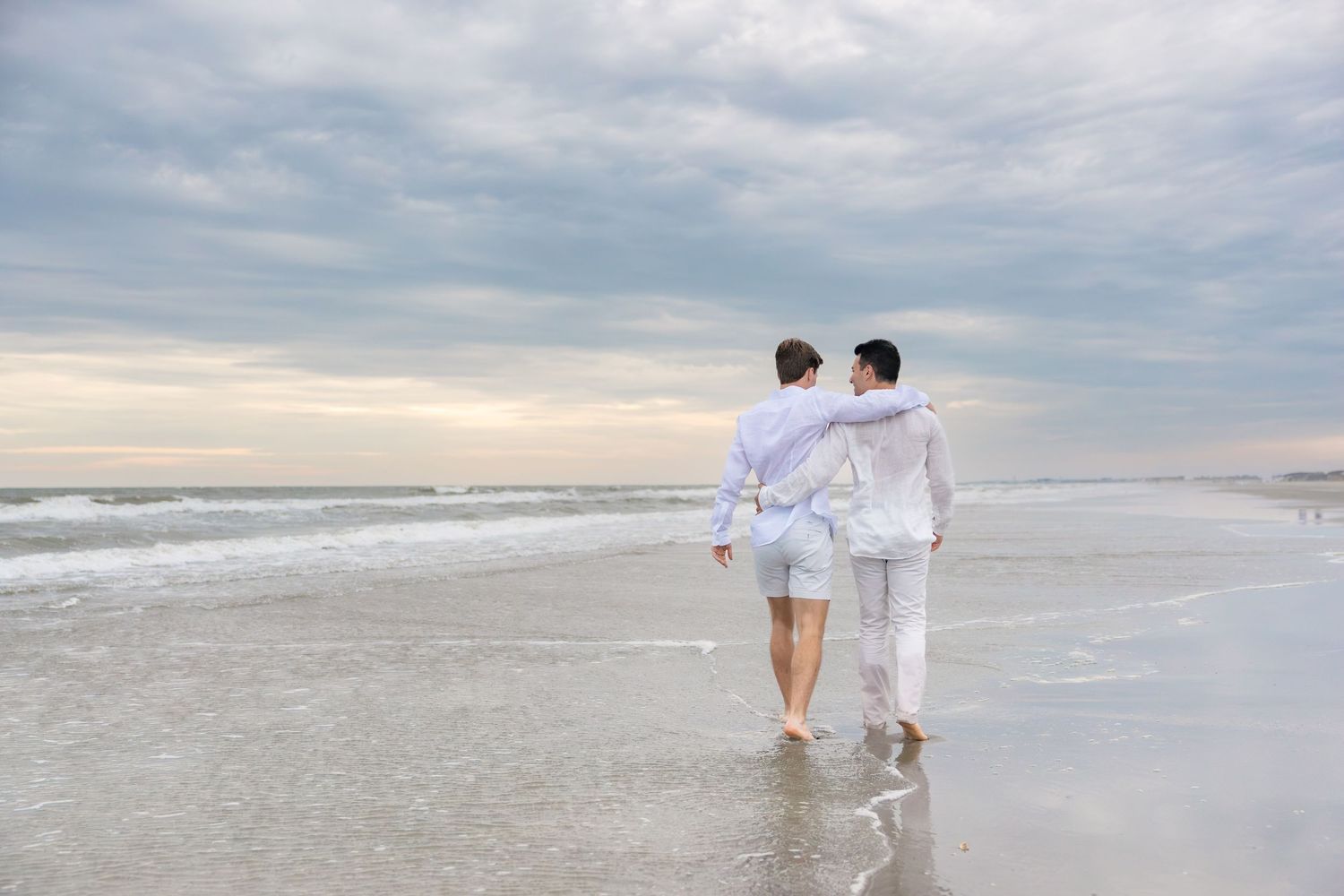 Two people in white outfits walking barefoot along a sandy beach shore with ocean waves and cloudy sky.