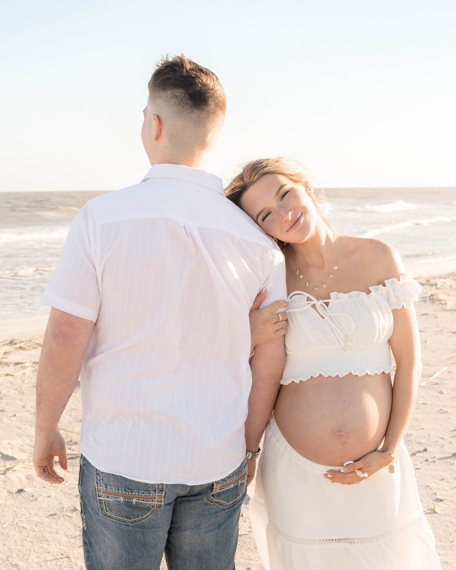 Pregnant couple in white outfits on beach, woman leaning on mans shoulder smiling, cradling baby bump.