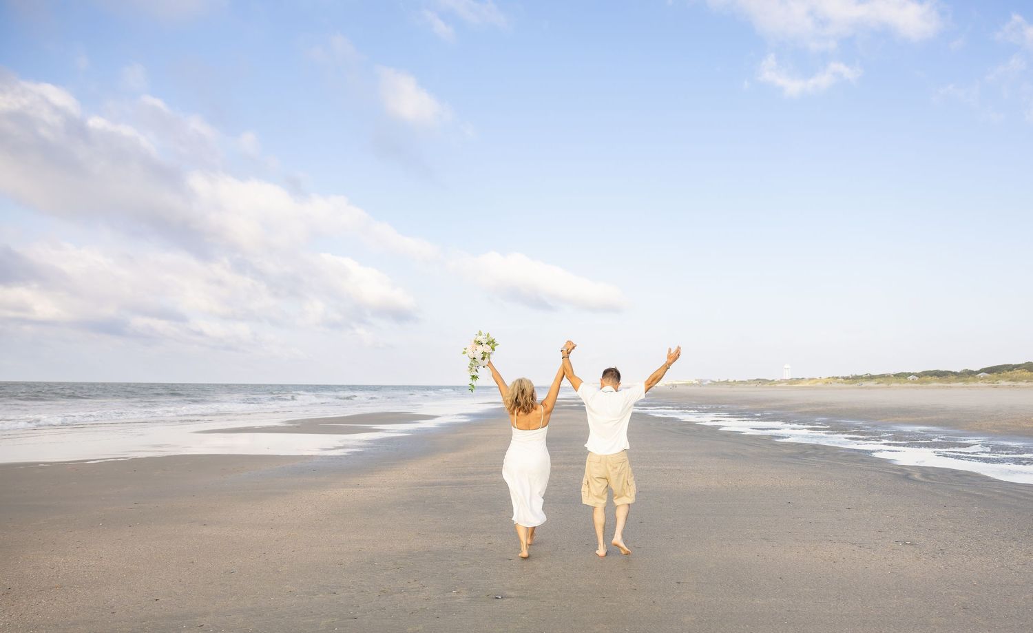Joyful newlywed couple celebrating on a sandy beach, raising their arms with a bouquet, ocean waves in the background.
