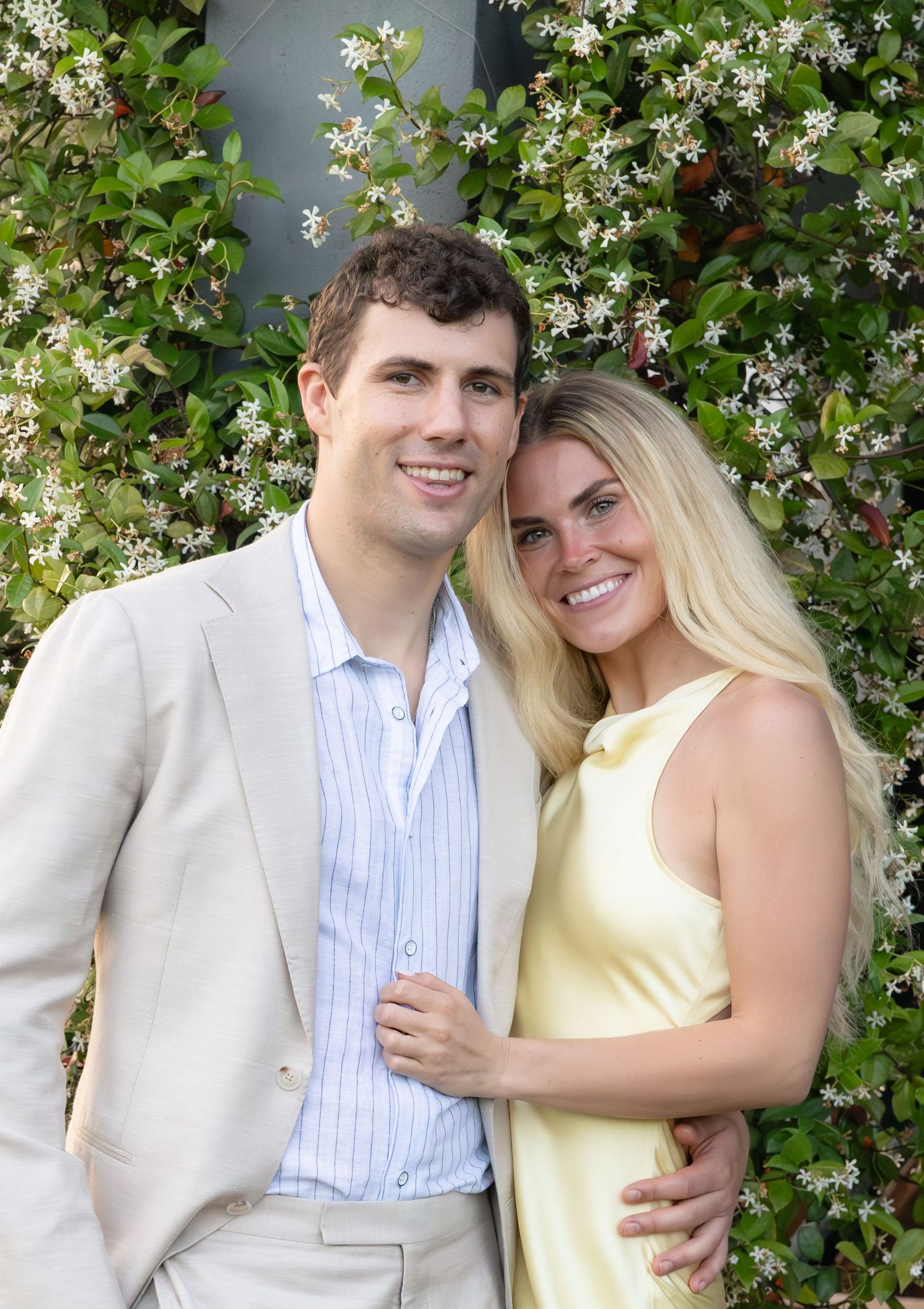 Smiling couple posing before flowering vine backdrop, man in beige suit, woman in yellow dress.