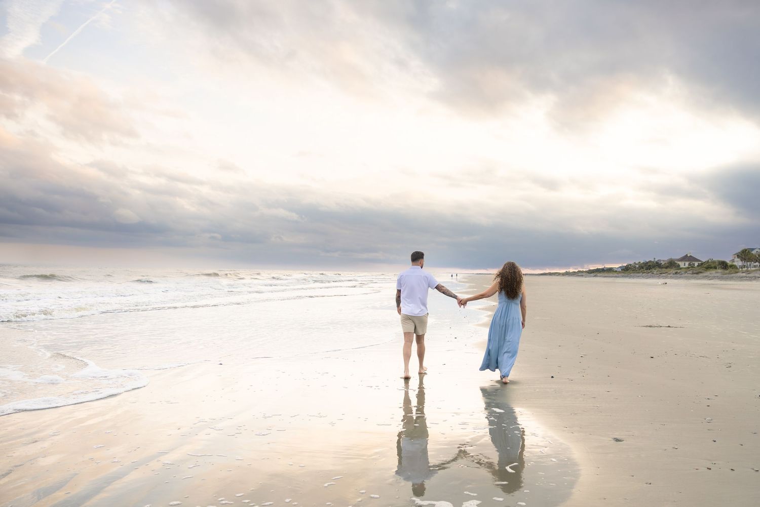 Couple holding hands walking along a serene beach at sunset with reflections on wet sand.
