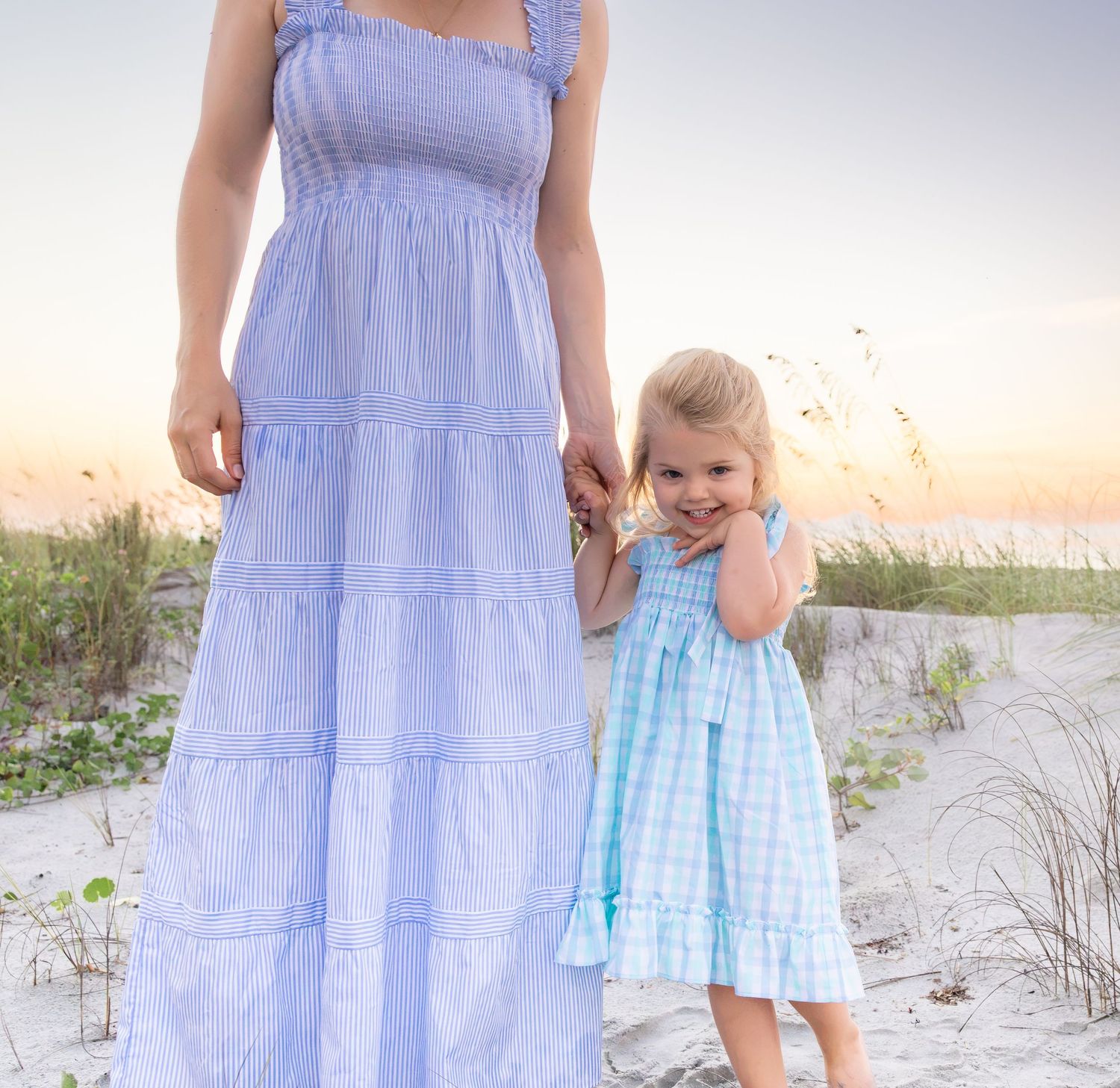 Mother and daughter in matching blue dresses holding hands on a sandy beach at sunset.