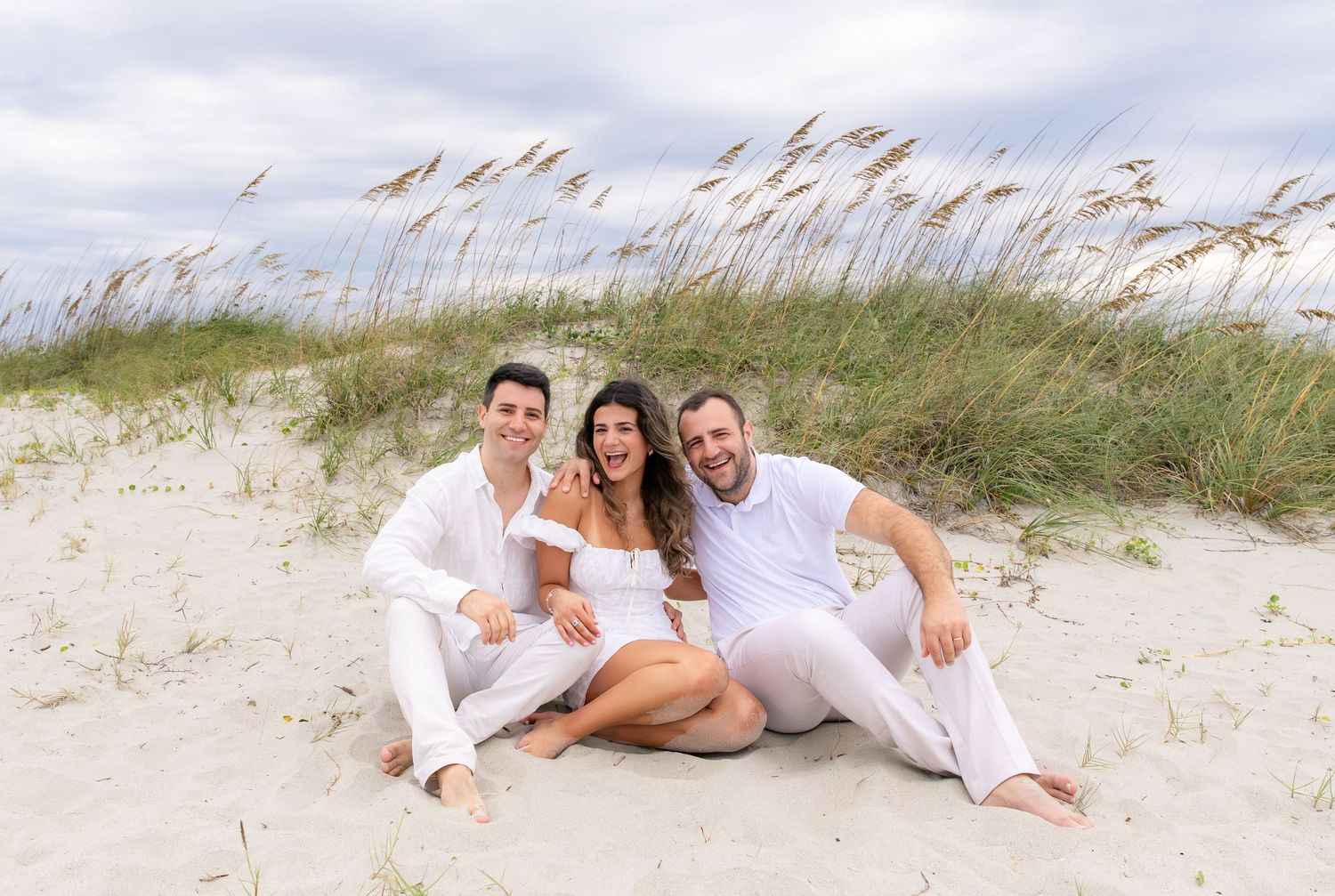 Three people in white outfits smiling together on a sandy beach with sea oats and cloudy sky backdrop.