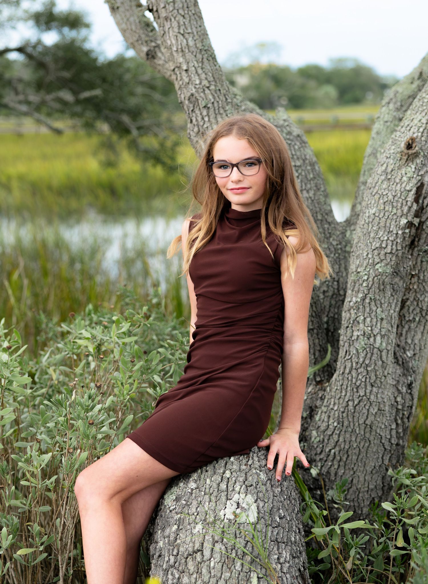 Young girl with long hair and glasses wearing a brown dress, sitting on a large tree trunk beside a marsh with tall green grass.