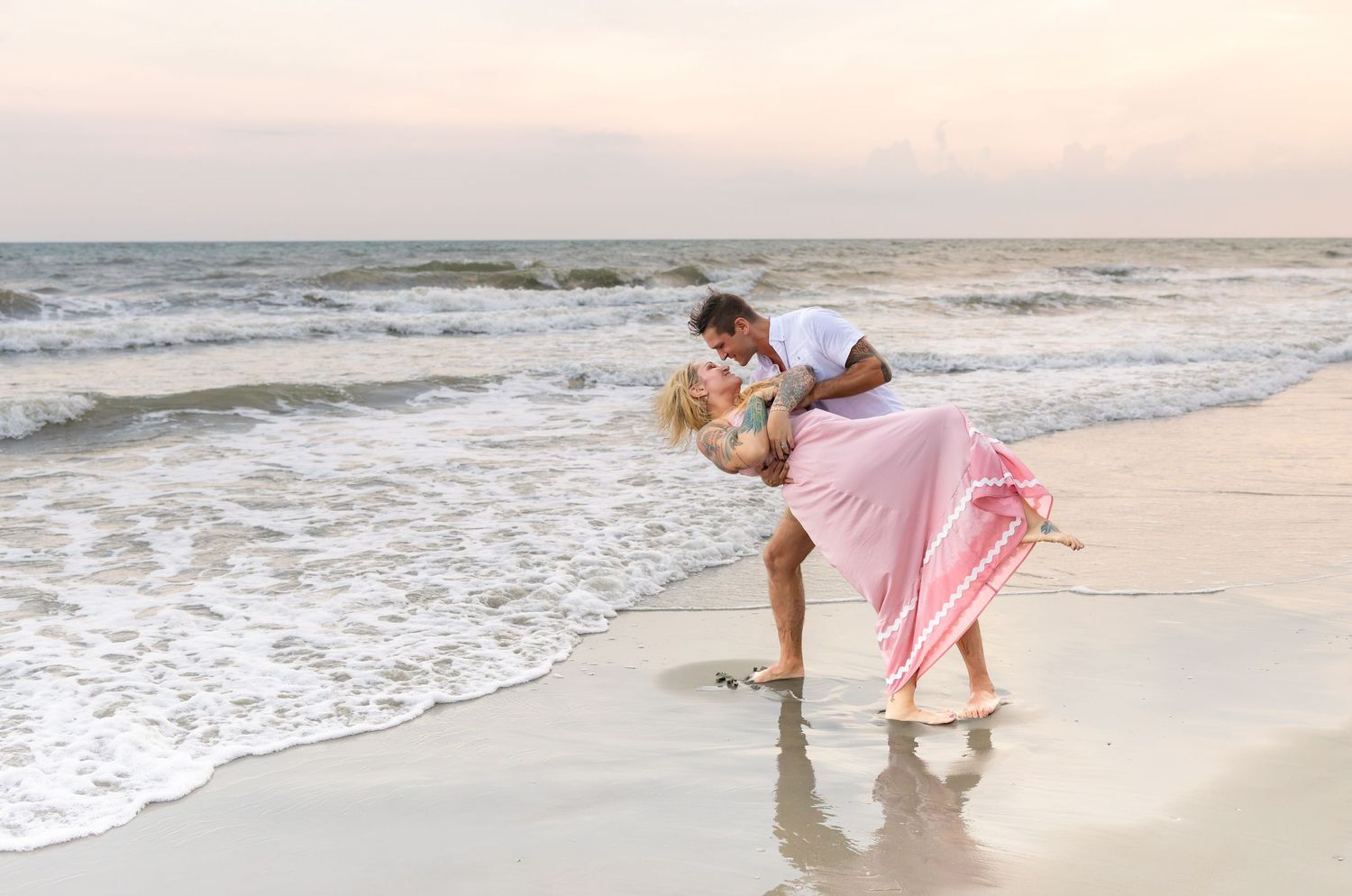 Couple in a romantic dip kiss on a sandy beach at sunset, the woman in a flowing pink skirt as ocean waves roll in behind them.