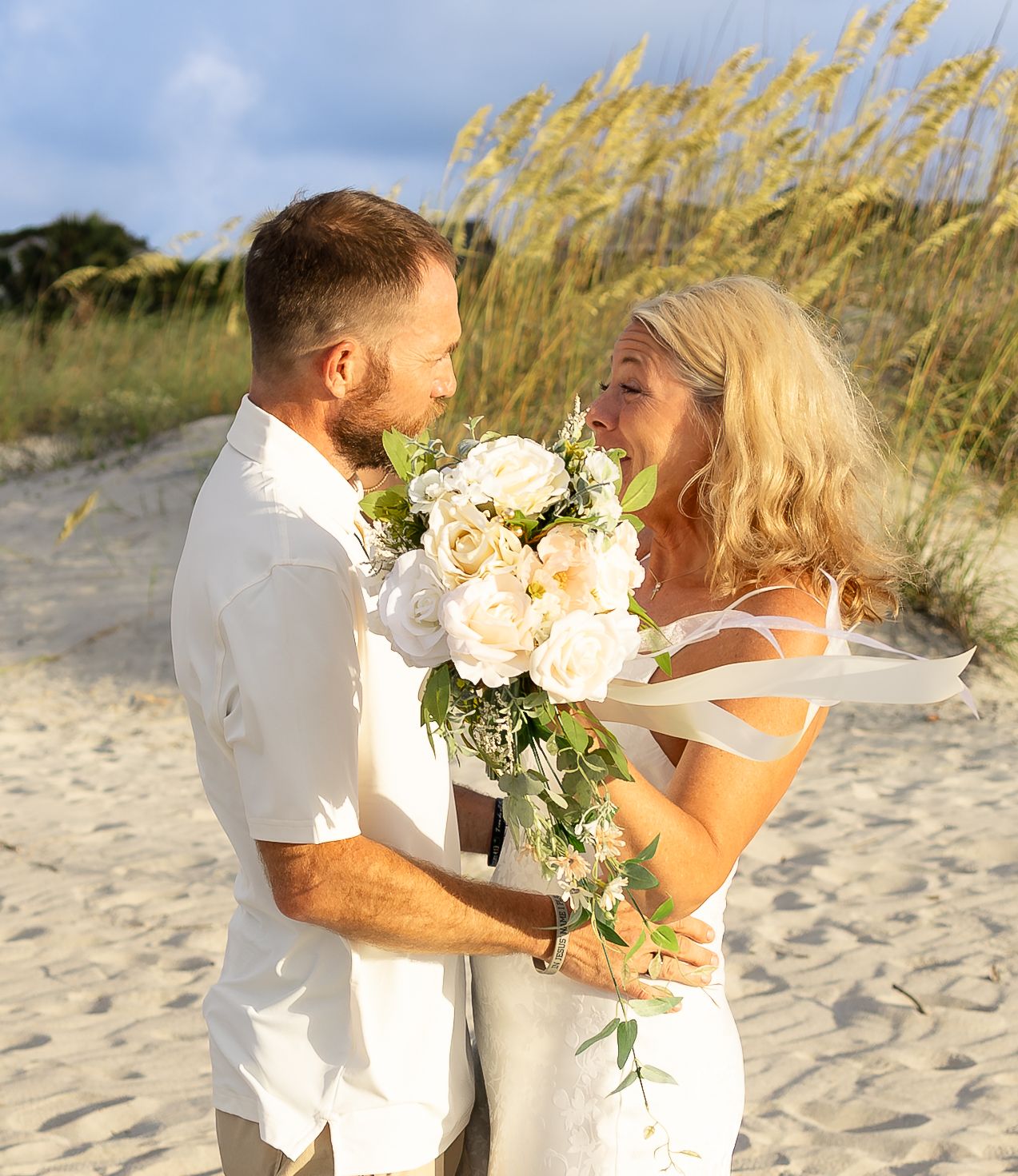 Couple in white attire sharing a romantic moment on a sandy beach, bride holding a white floral bouquet.