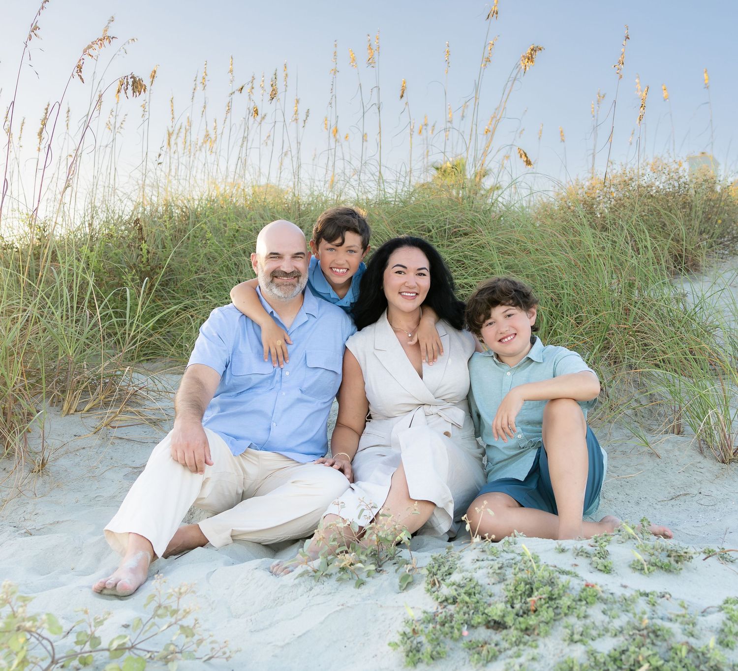 Happy family of four sitting together on sandy beach dunes with sea oats, smiling for a portrait photo.