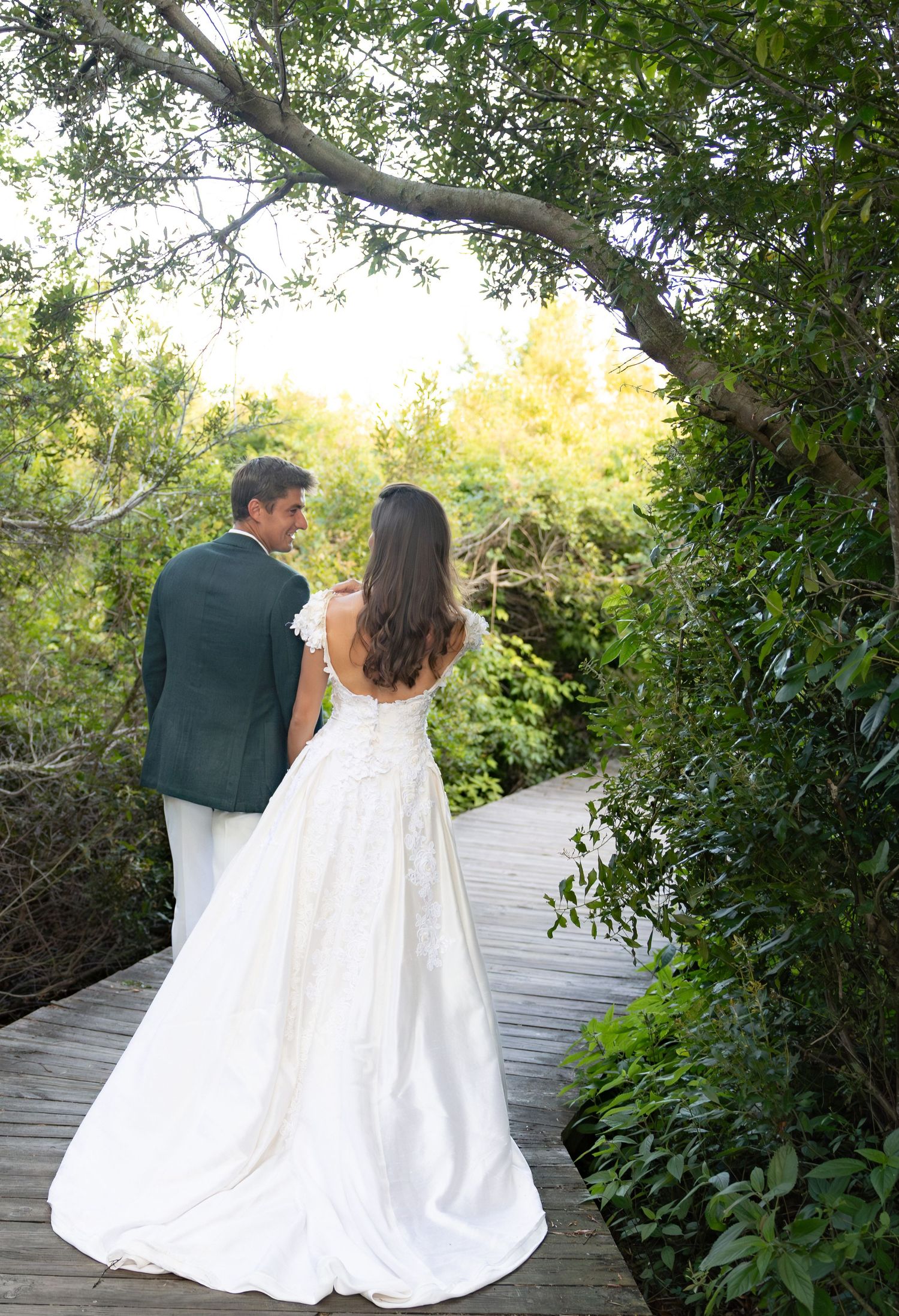 Bride and groom standing on wooden boardwalk surrounded by lush green trees in romantic outdoor wedding photo.