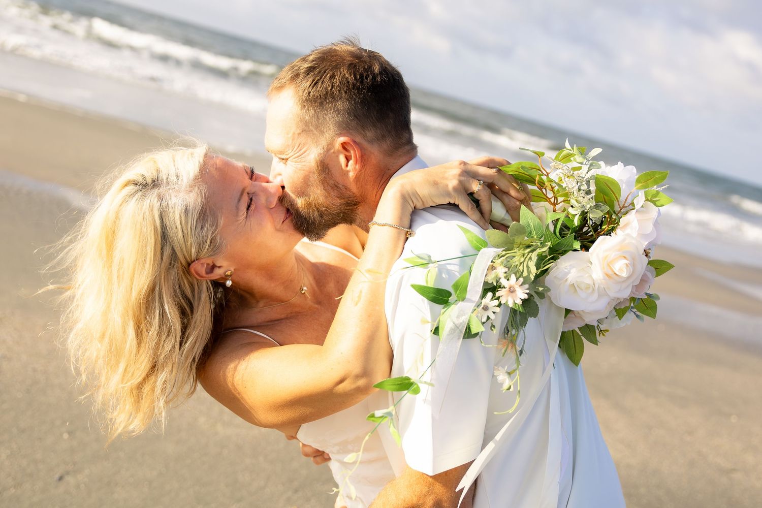 A couple shares a romantic kiss on a beach wedding, bride holding white floral bouquet with greenery.