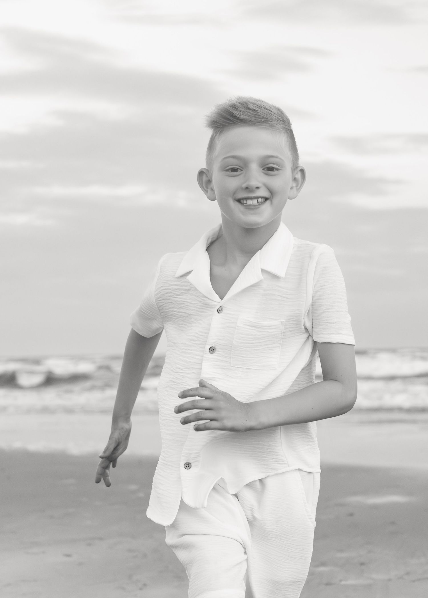 Smiling boy in white outfit running on the beach in a black and white photo.
