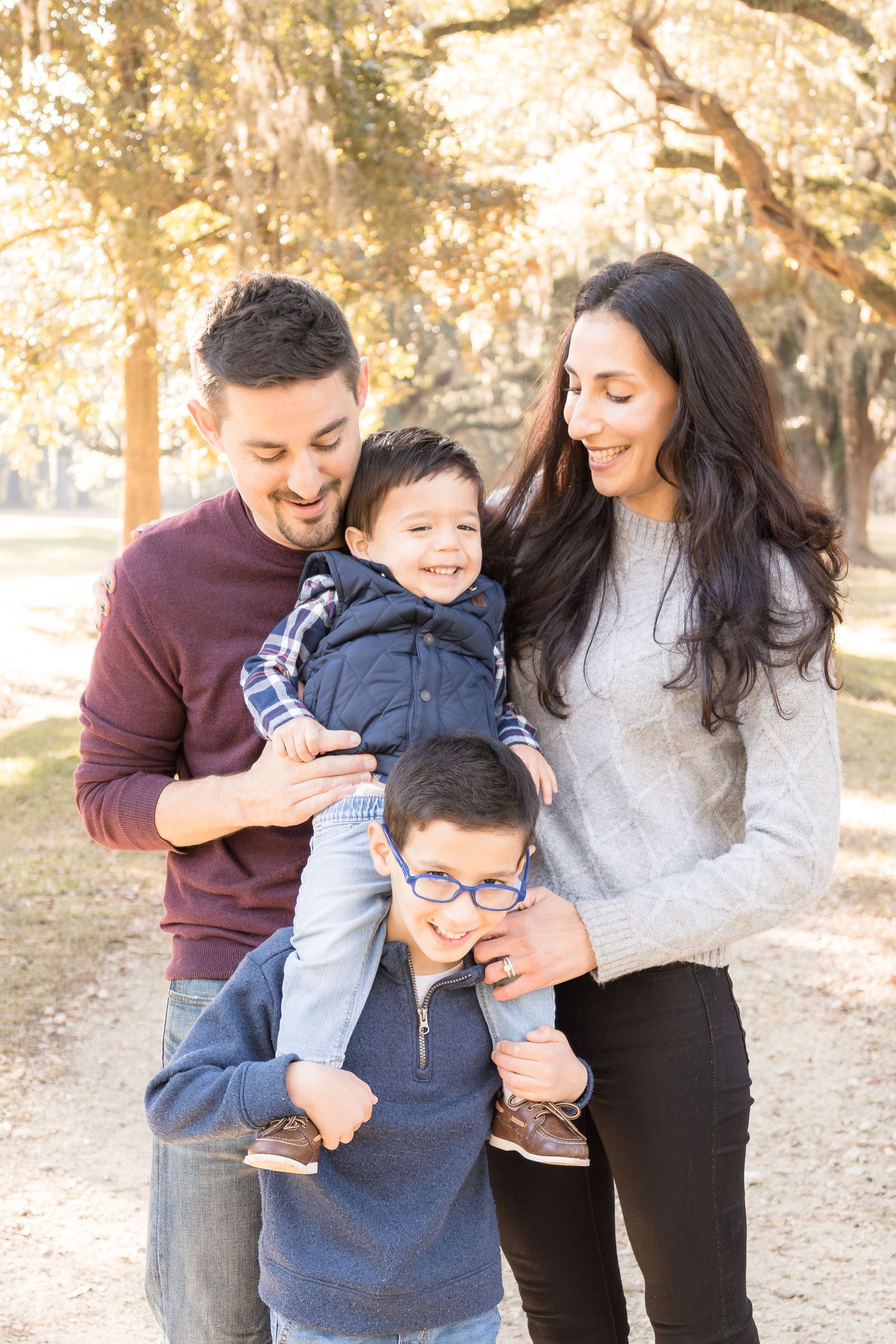 Smiling family of four posing outdoors under oak trees, with parents embracing two happy young boys during a sunny autumn portrait session.