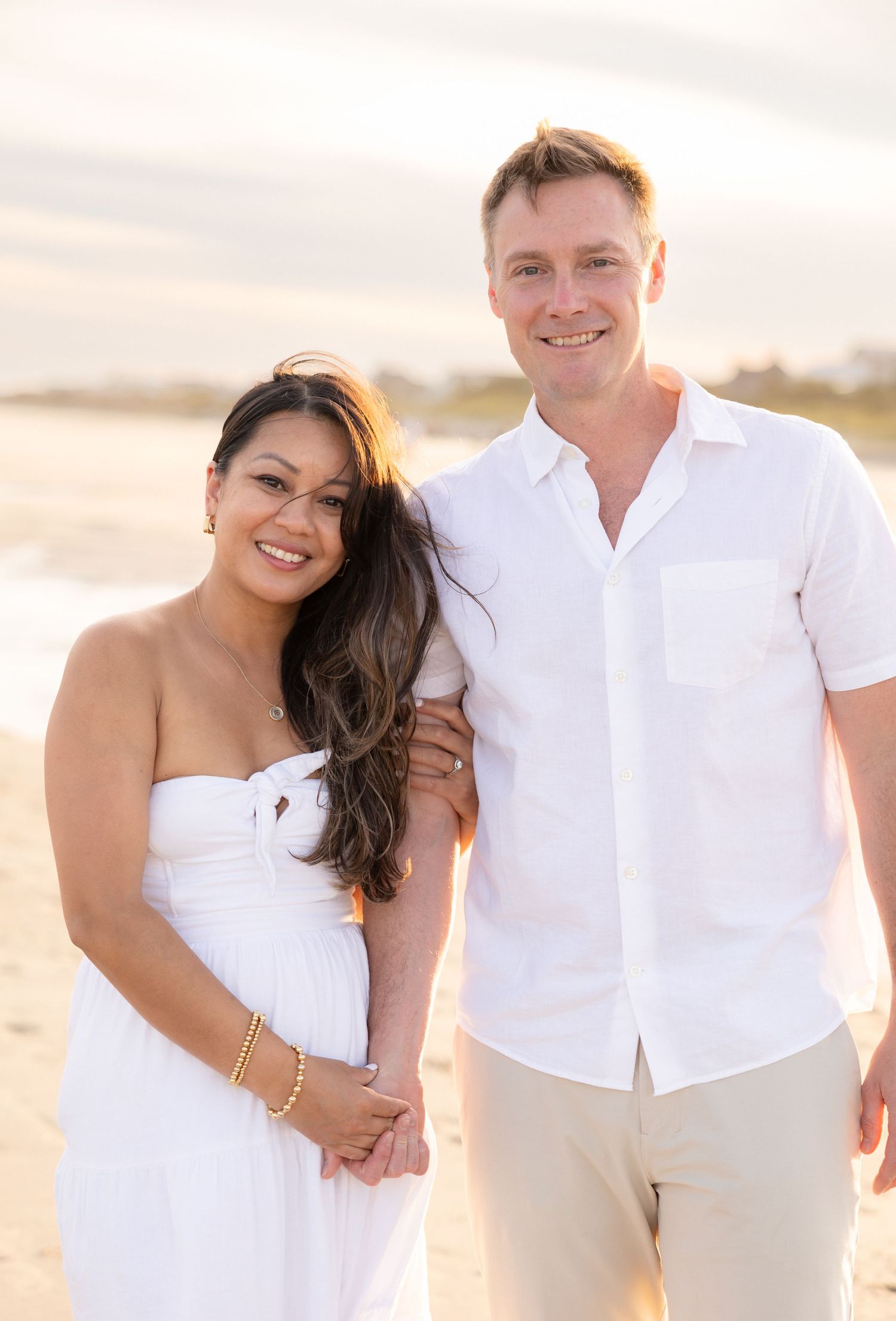 Couple in white outfits posing on a beach at sunset, woman in strapless dress and man in button-up shirt smiling.