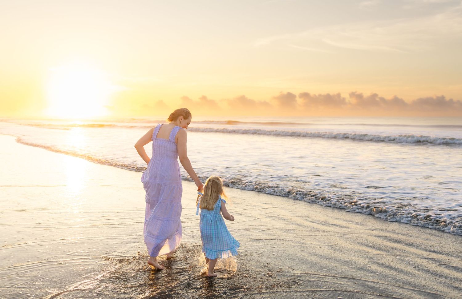 Mother and daughter walking on a sunny beach at golden sunset, wearing blue dresses by the ocean waves.