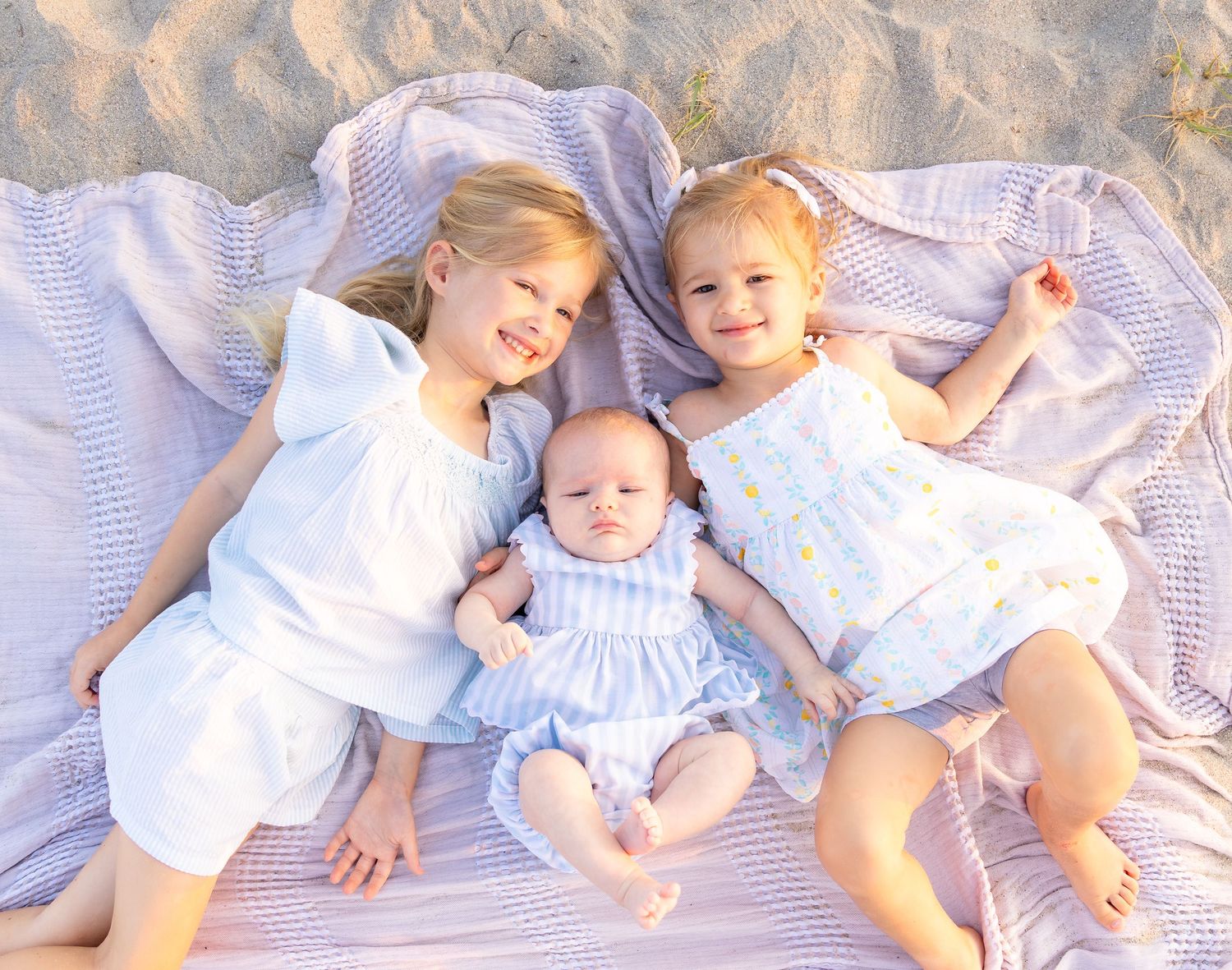 Three young sisters lying on a blanket at the beach, two toddlers flanking a smiling baby in blue.