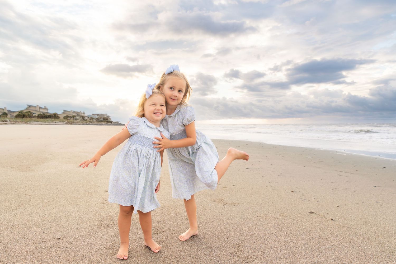 Two young girls in matching blue dresses laugh and pose together on a sandy beach with ocean waves behind them.