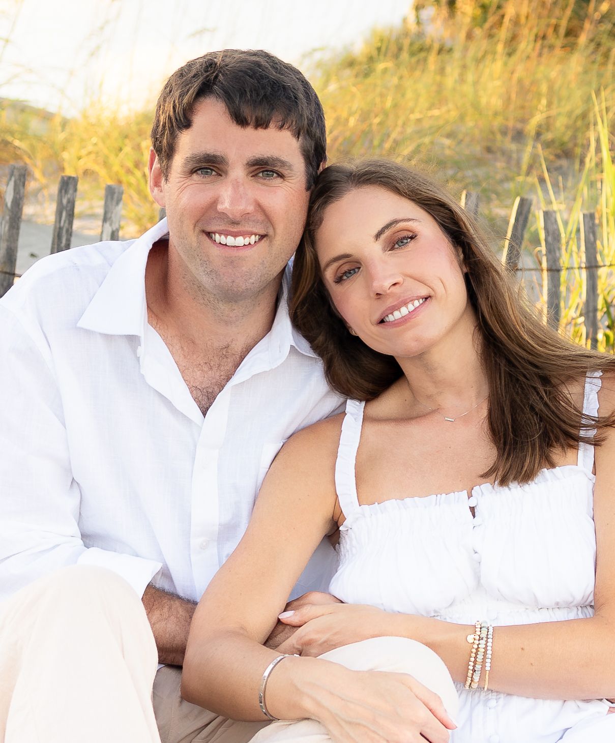Smiling couple in white outfits posing together on a sunny beach with sand dunes in the background.