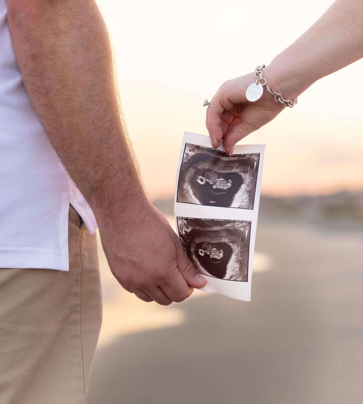 Couple holding hands and ultrasound photos at sunset, announcing pregnancy with sonogram pictures outdoors.