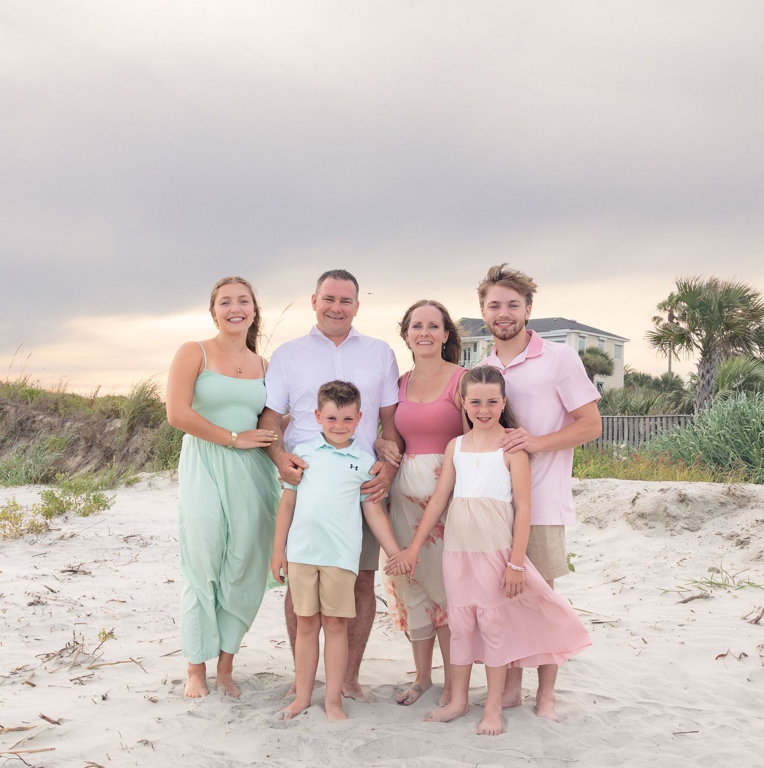 Family of six posing together on a sandy beach at sunset, wearing coordinated pastel outfits.