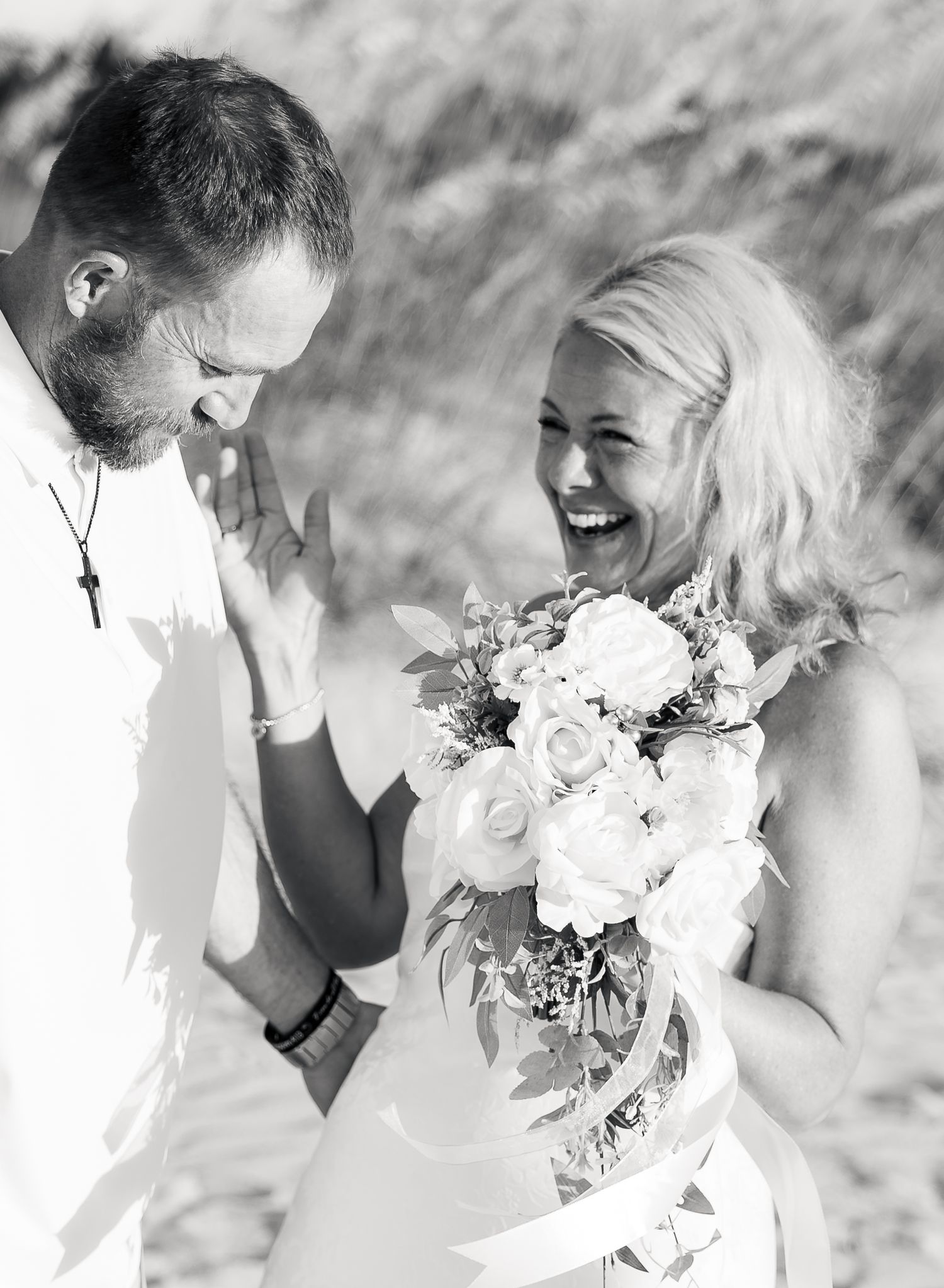 Black and white photo of a laughing bride holding a white rose bouquet with her groom on a sandy beach.