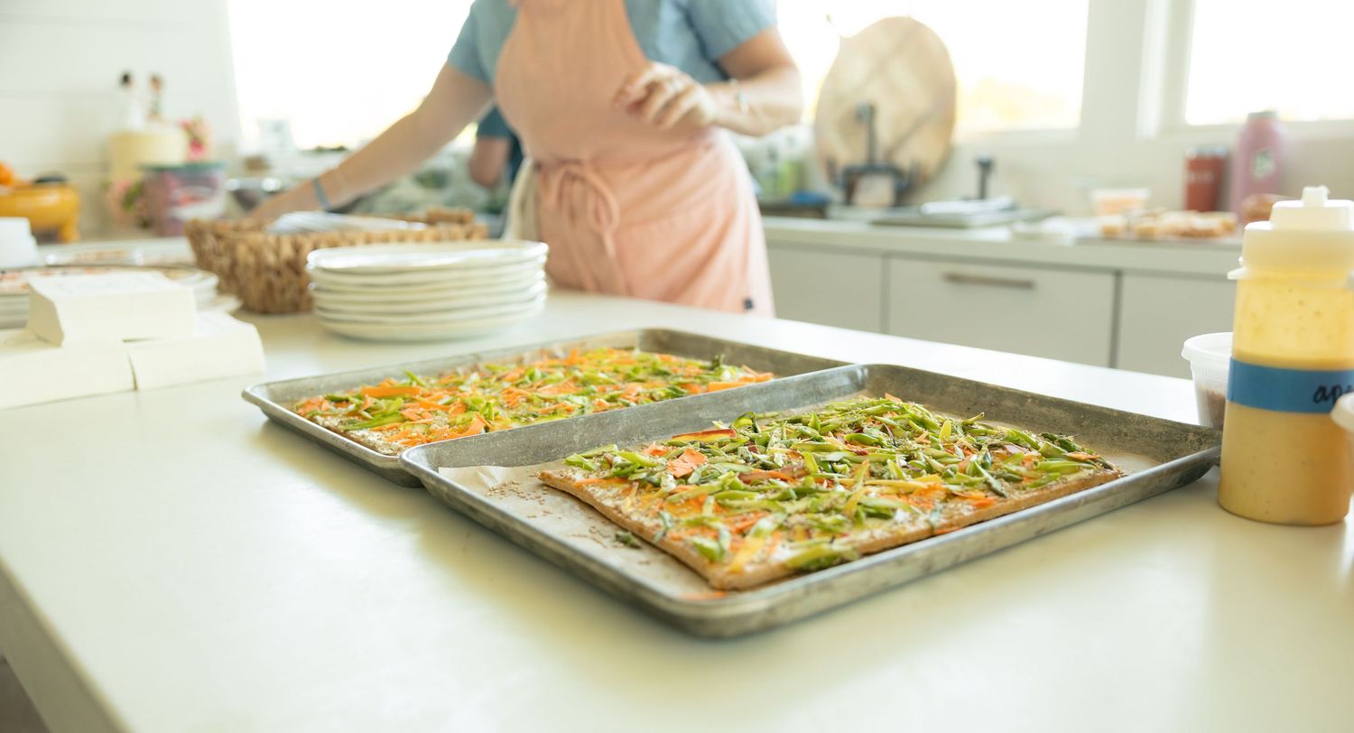 Two freshly baked flatbread pizzas topped with vegetables on baking sheets on a bright kitchen counter.