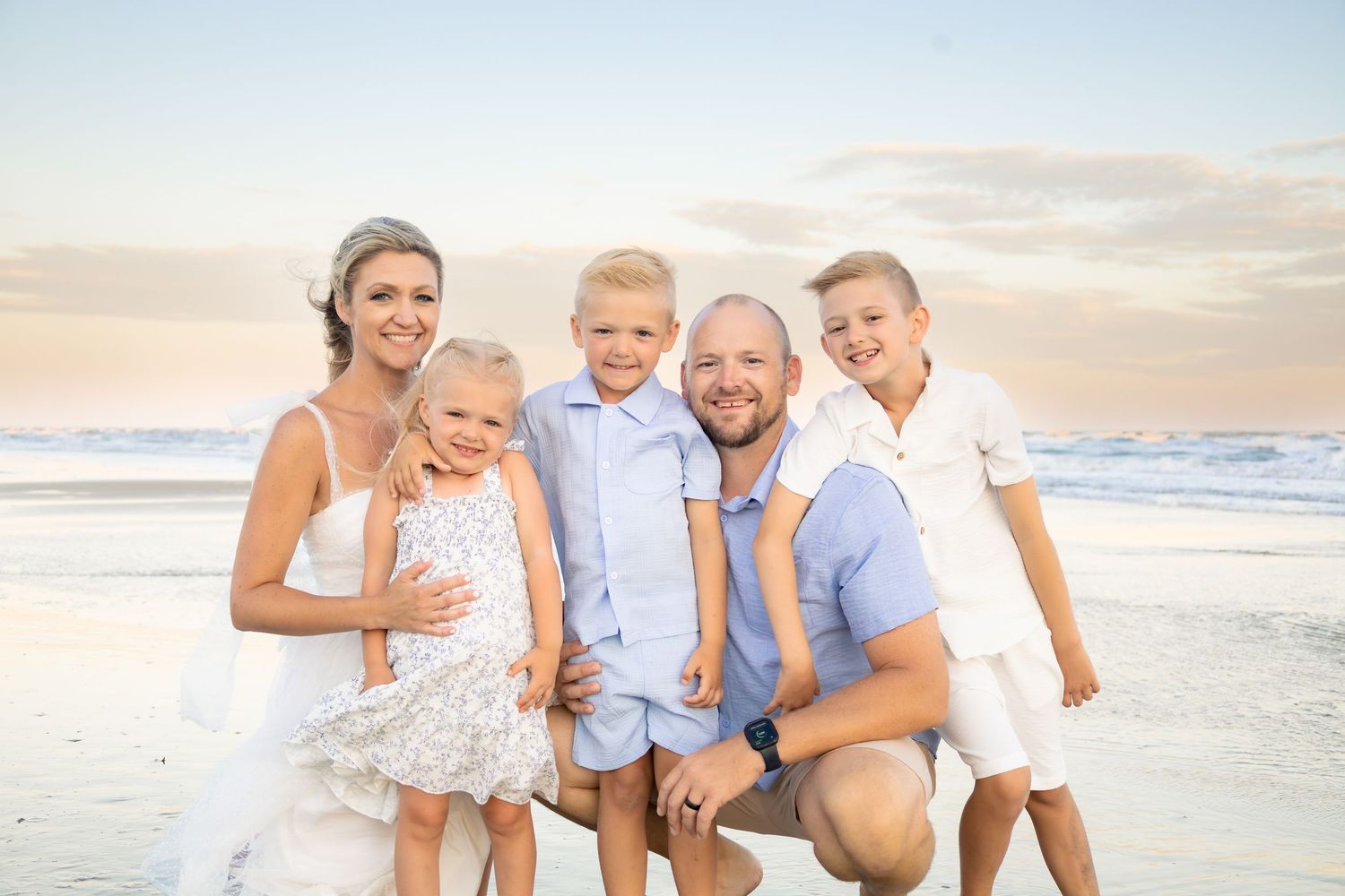 A happy family of five poses together on a beach at sunset, smiling for a portrait photo.