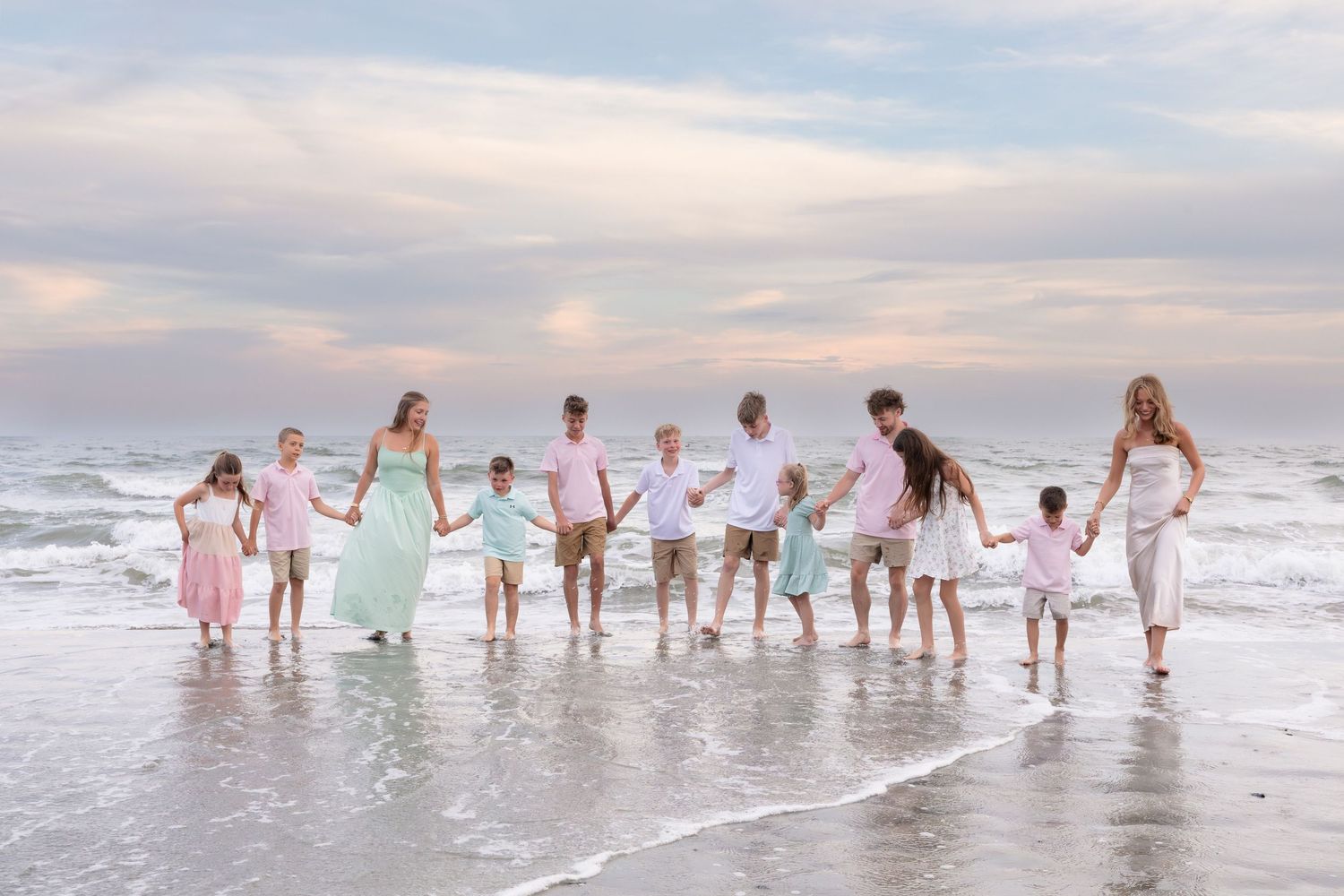 Large family holding hands walking along beach shoreline at sunset in pastel pink and mint outfits.
