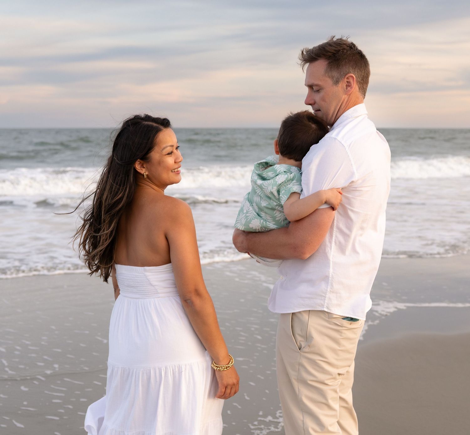 A young family stands on a beach at sunset, with a man holding a baby and a woman in a white dress smiling back.