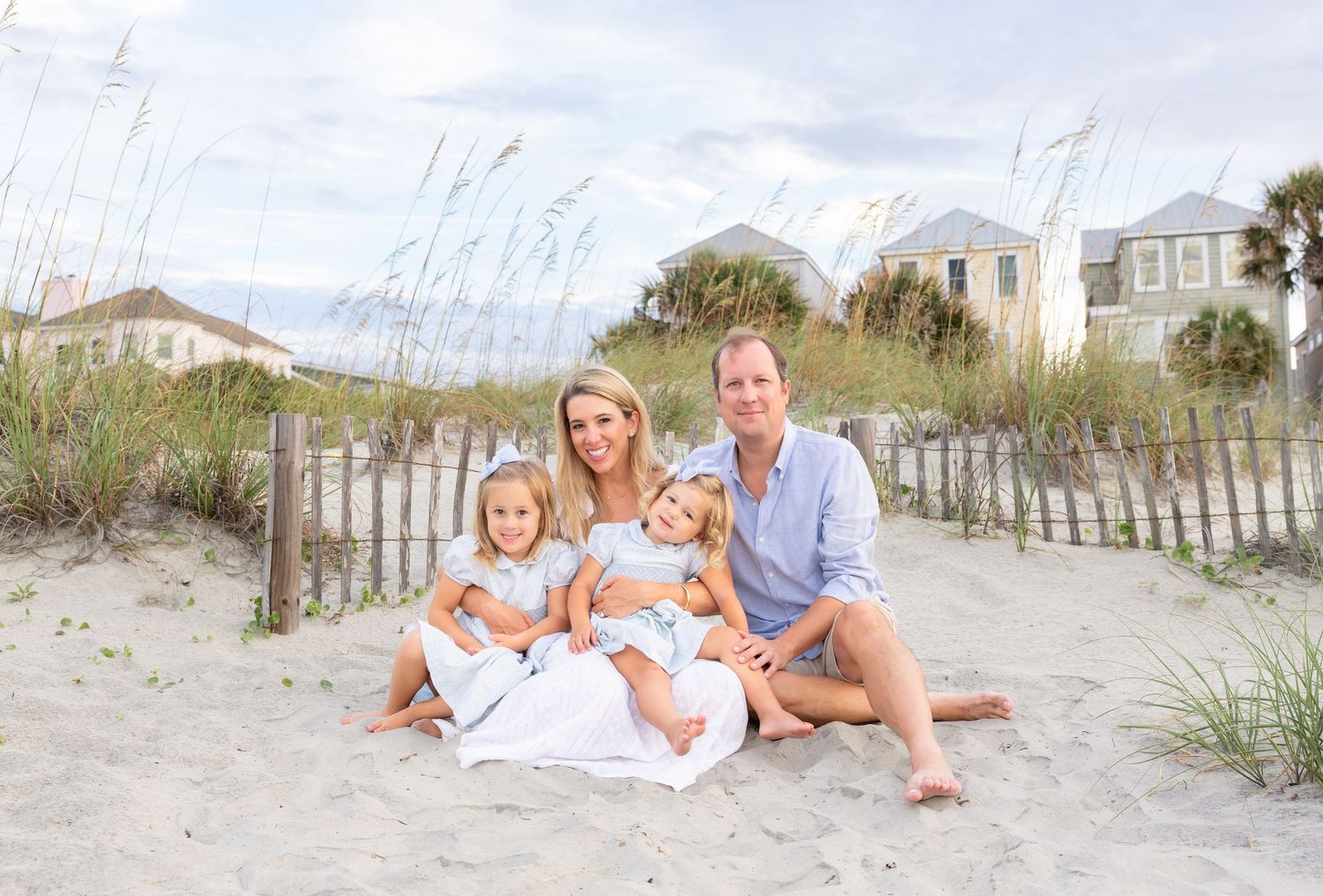 Family of four poses on sandy beach dunes with sea oats and pastel beach houses in background on sunny day.